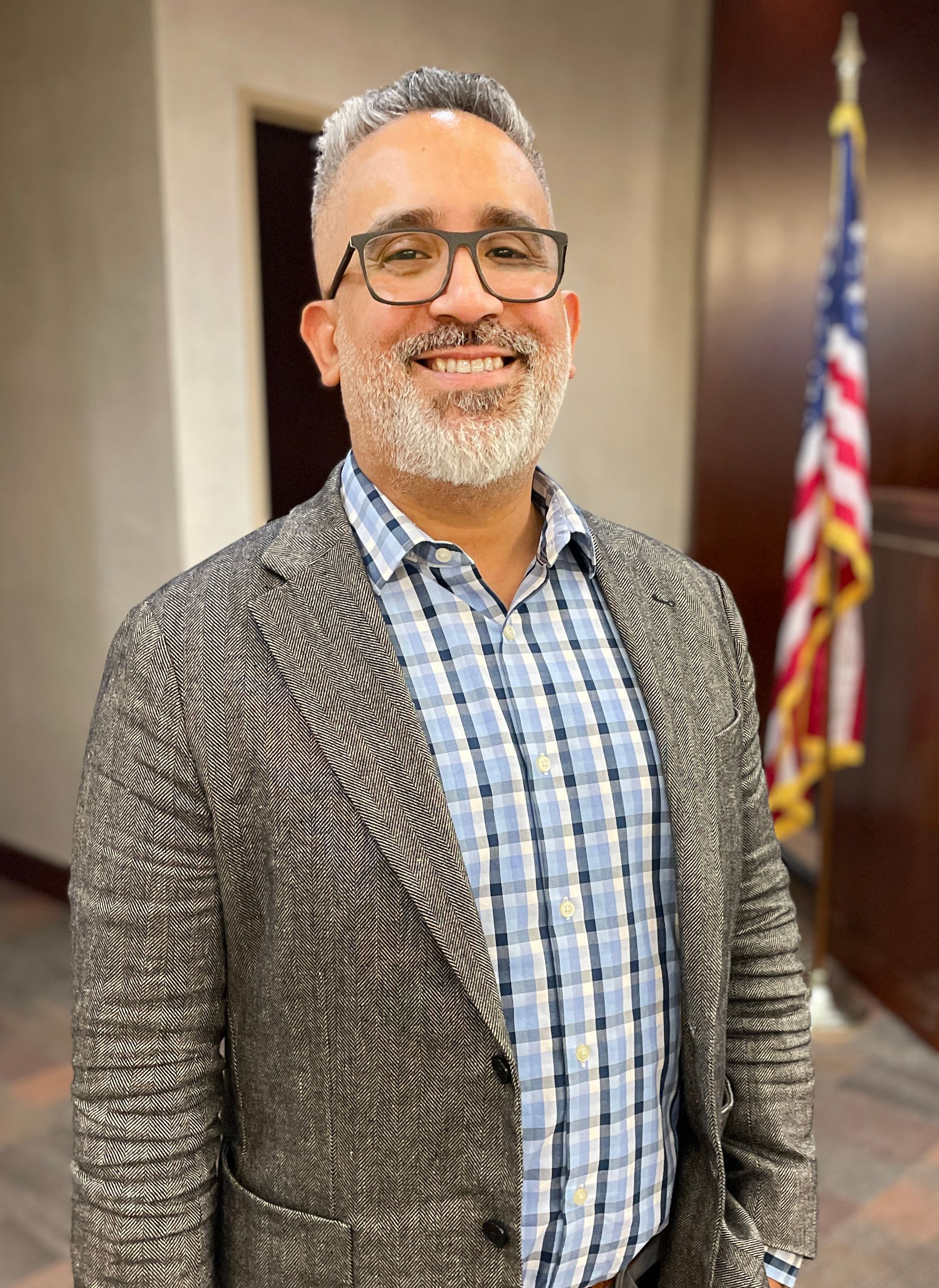 A man with glasses and a beard is standing in front of an american flag.