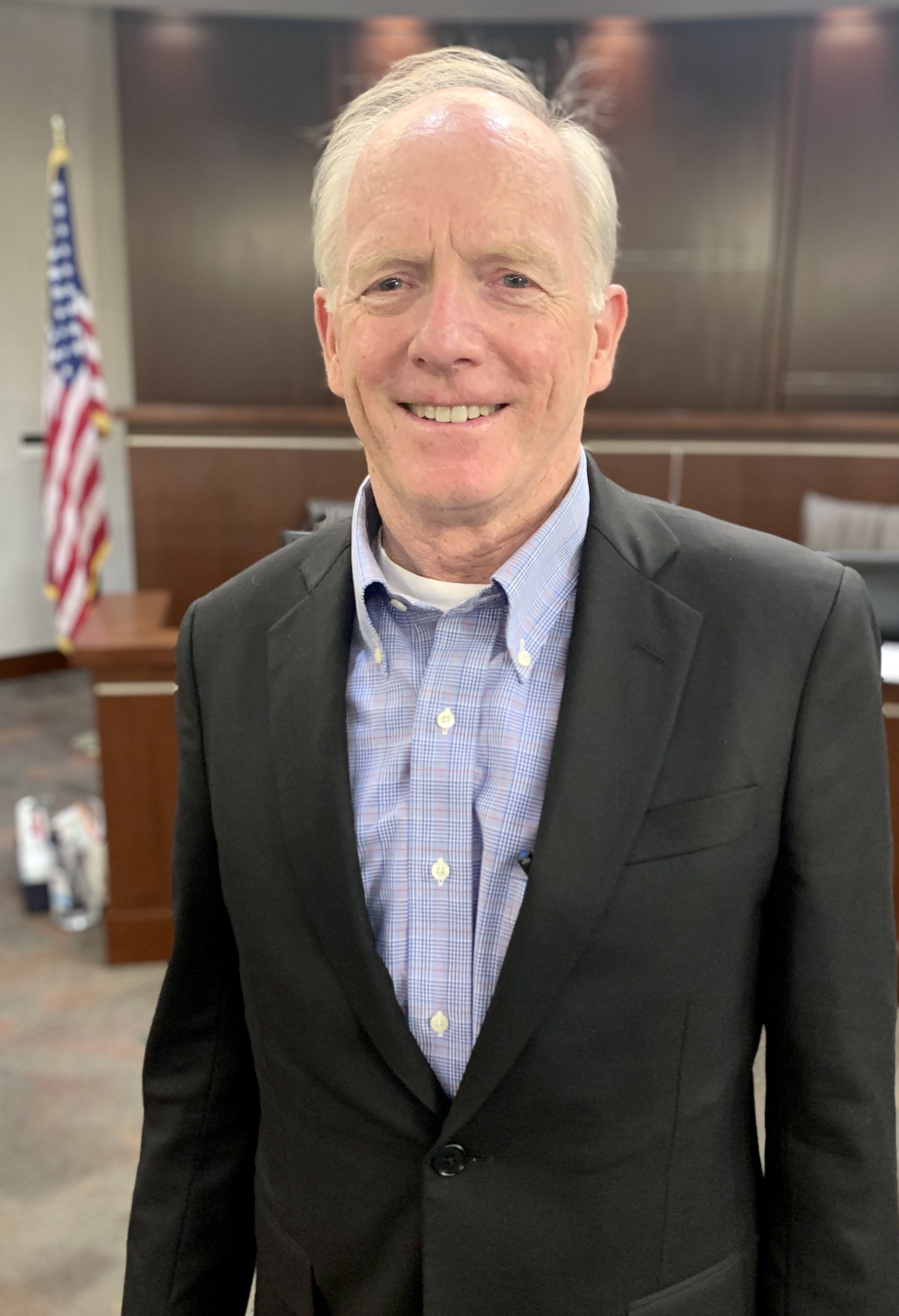 A man in a suit and blue shirt is standing in front of an american flag.