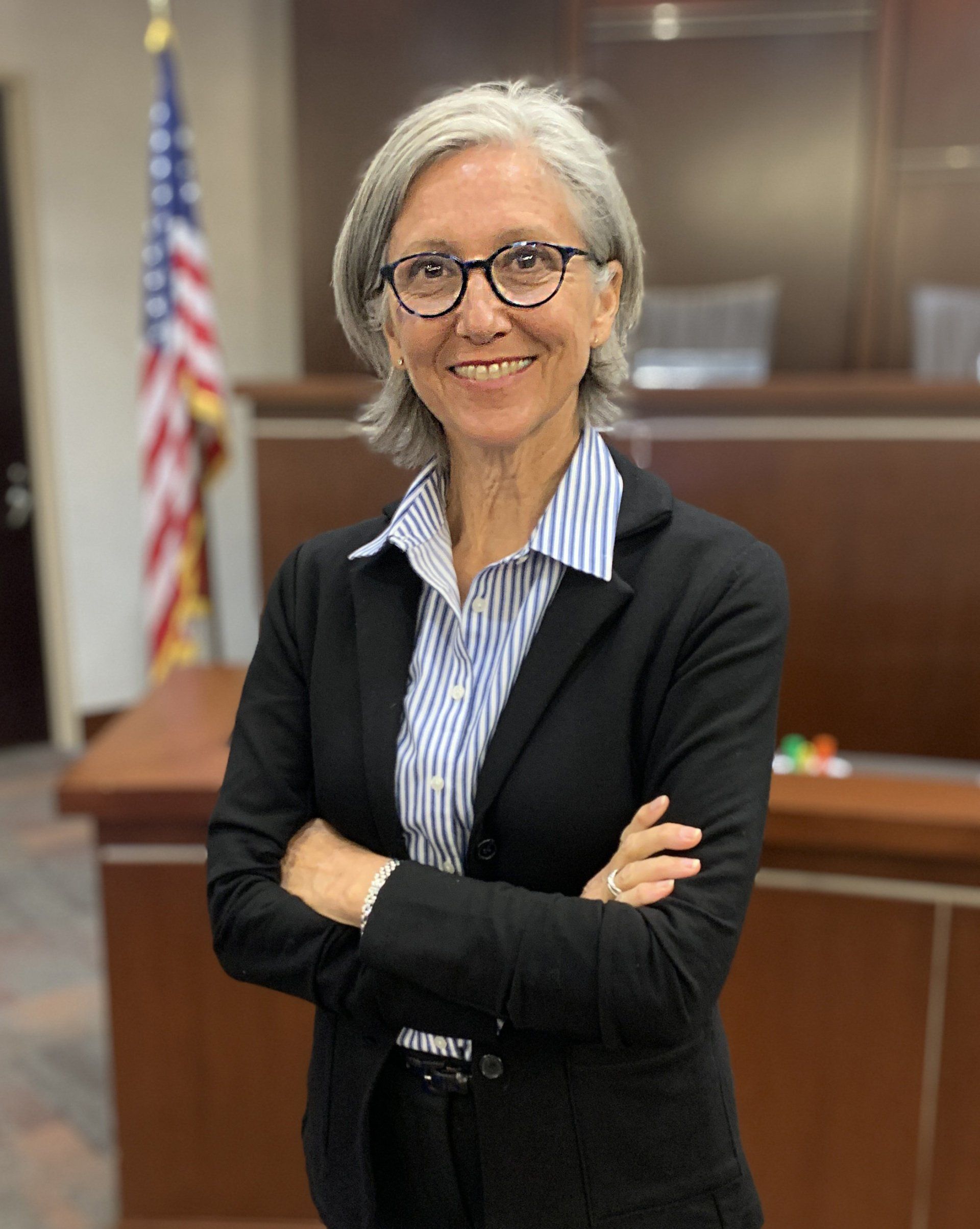 A woman wearing glasses and a black jacket is standing in front of an american flag.