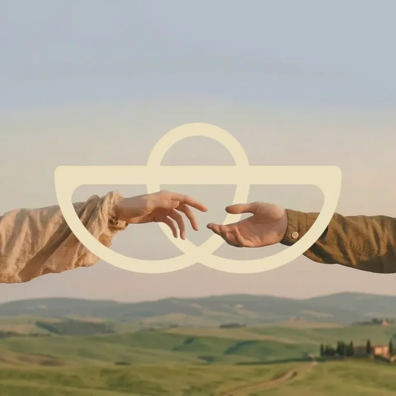 Two hands reaching toward each other over a soft, cream-colored symbol above rolling green hills