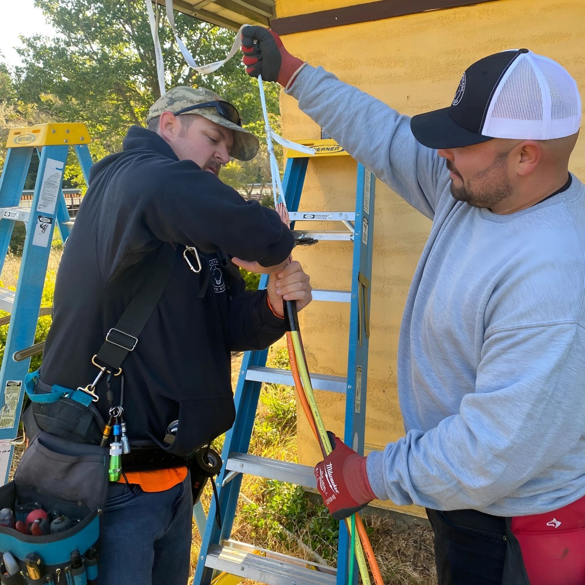 The Electric Pro - Two men are working on a ladder with one wearing a hat that says ny
