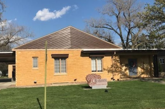 Yellow brick building with a brown metal roof, windows, and a stone monument on the lawn.