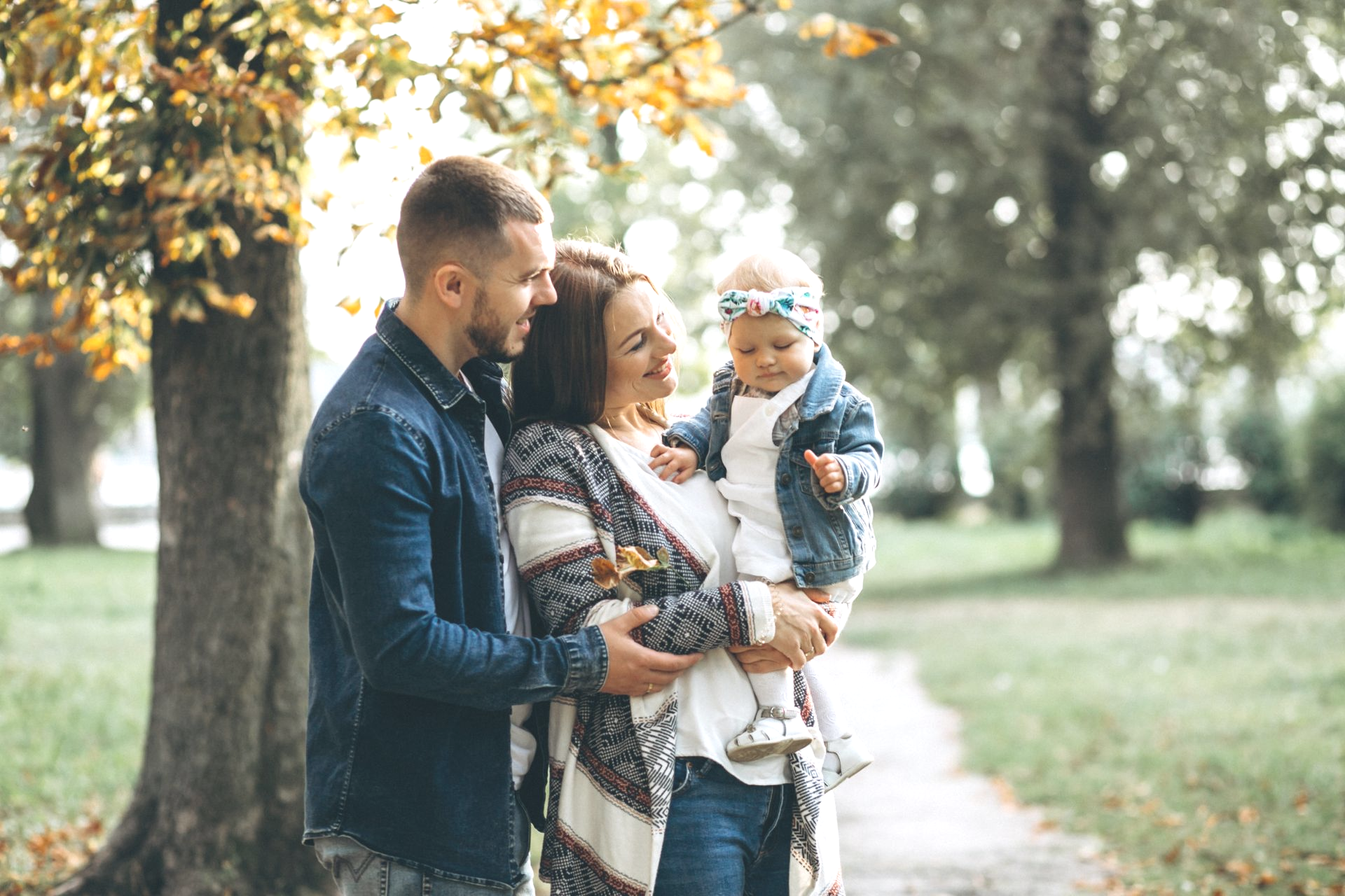 Family of three in a park; father, mother holding baby, all wearing casual clothing. Autumn leaves, path in background.