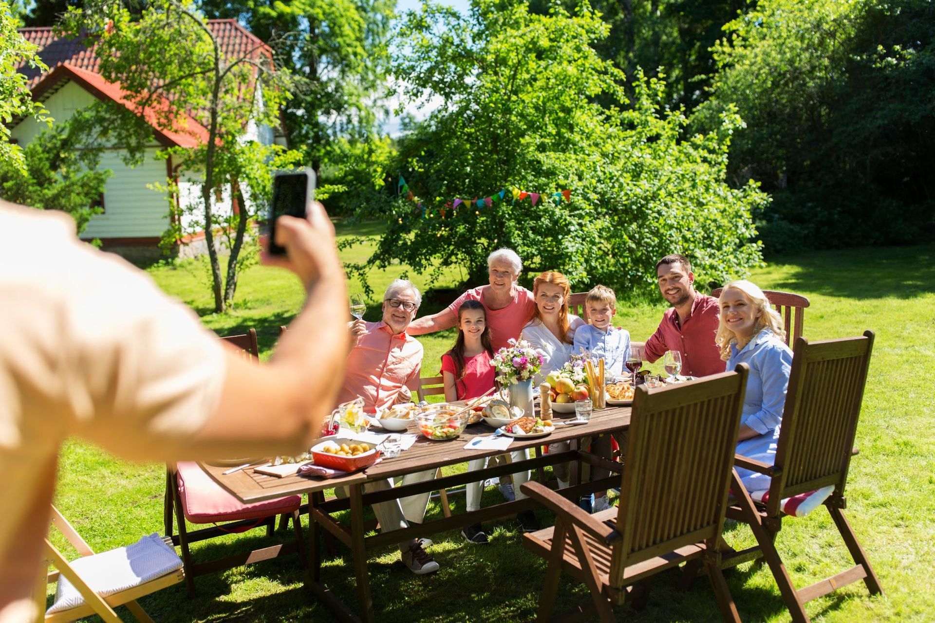 Family taking a photo at an outdoor table laden with food; sunny day in a backyard.