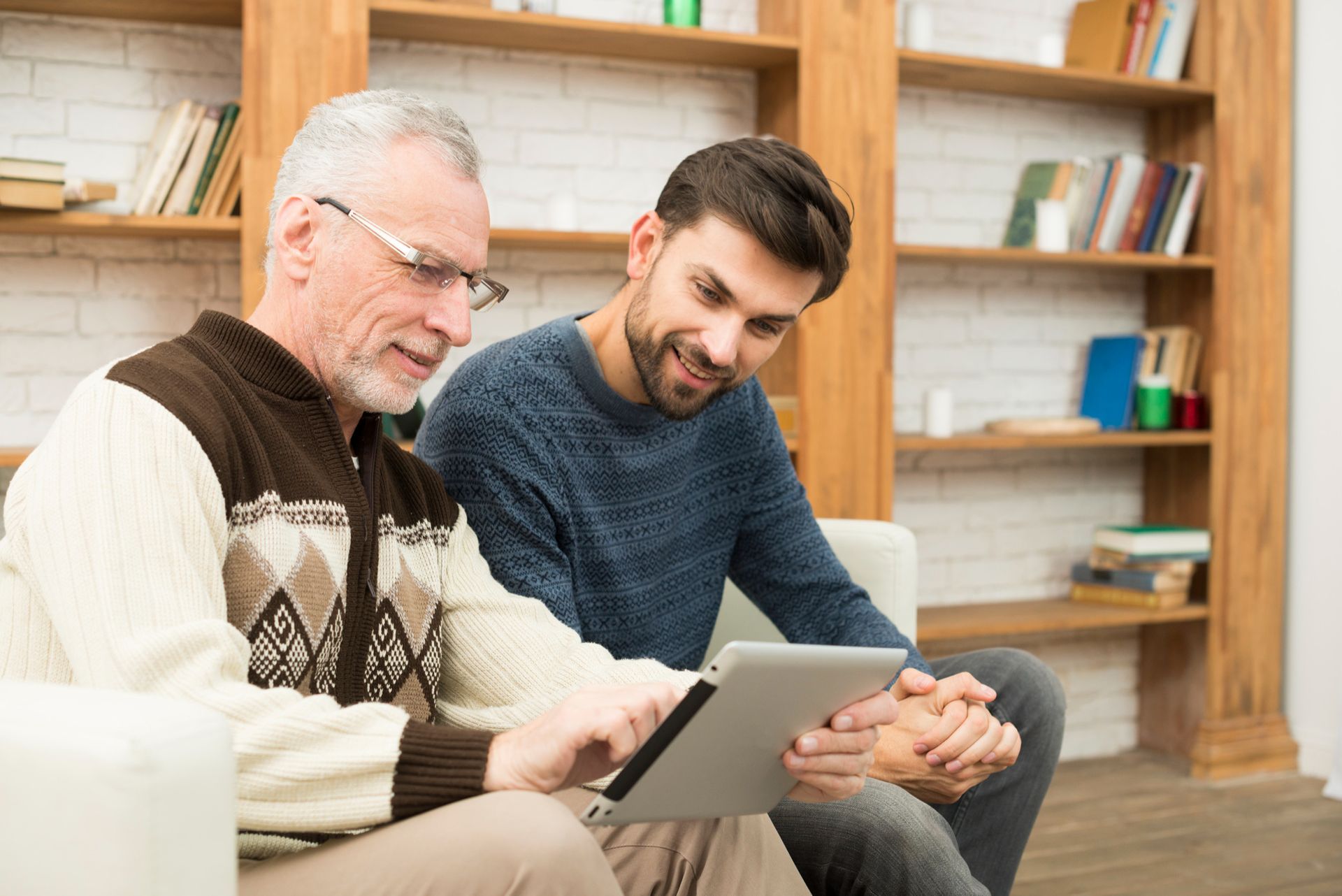 Older man with glasses and younger man looking at a tablet together, smiling. They are sitting indoors near a bookshelf.