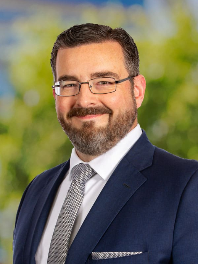 Man with glasses, beard, and suit smiling at the camera; green and blue background.