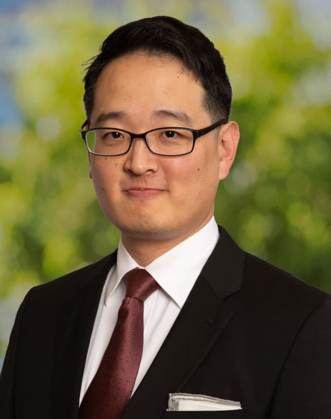 Man in suit and glasses, looking at the camera, with a maroon tie, and a green and blue background.