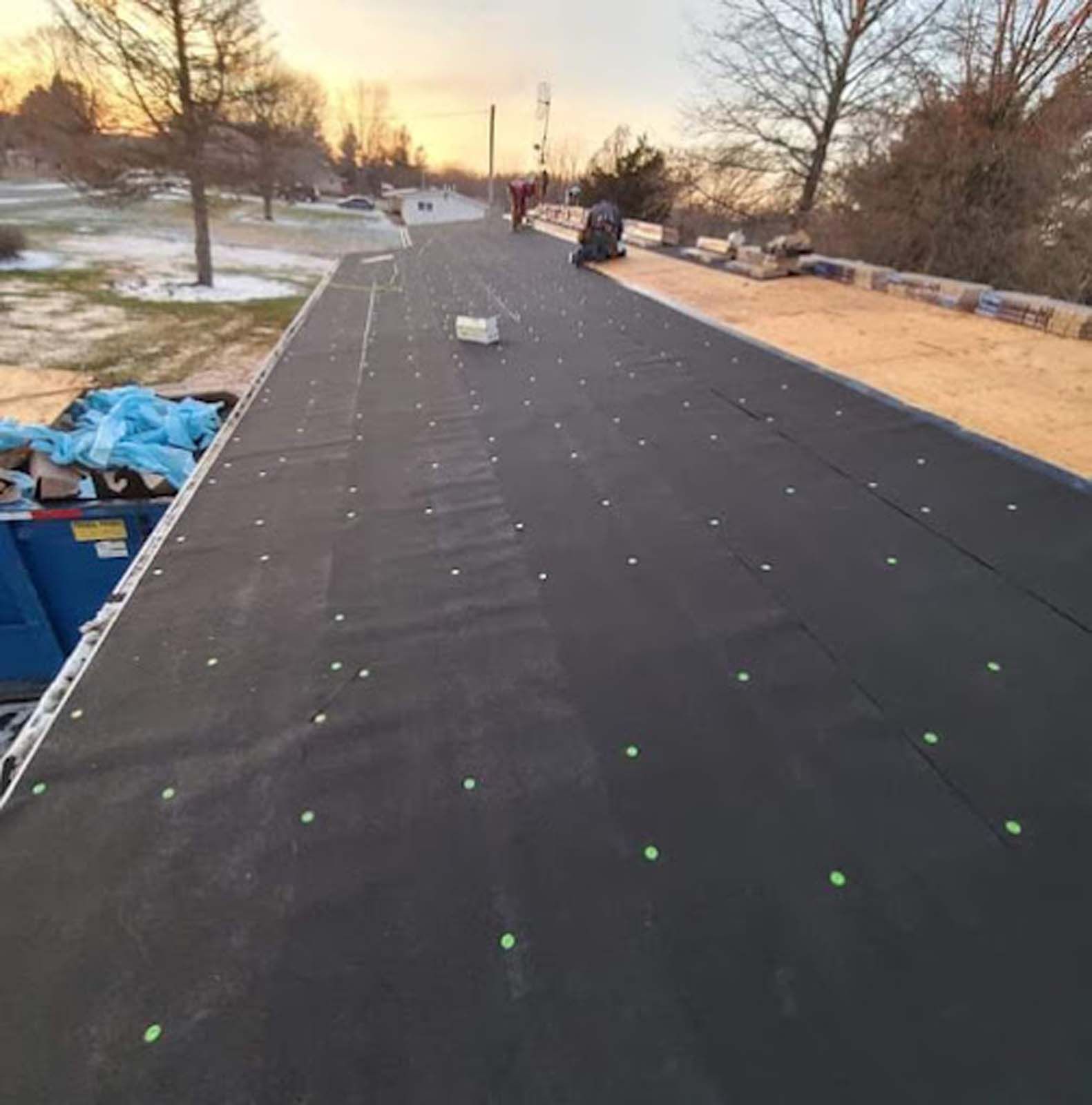 A person is working on a roof with a dumpster in the background.