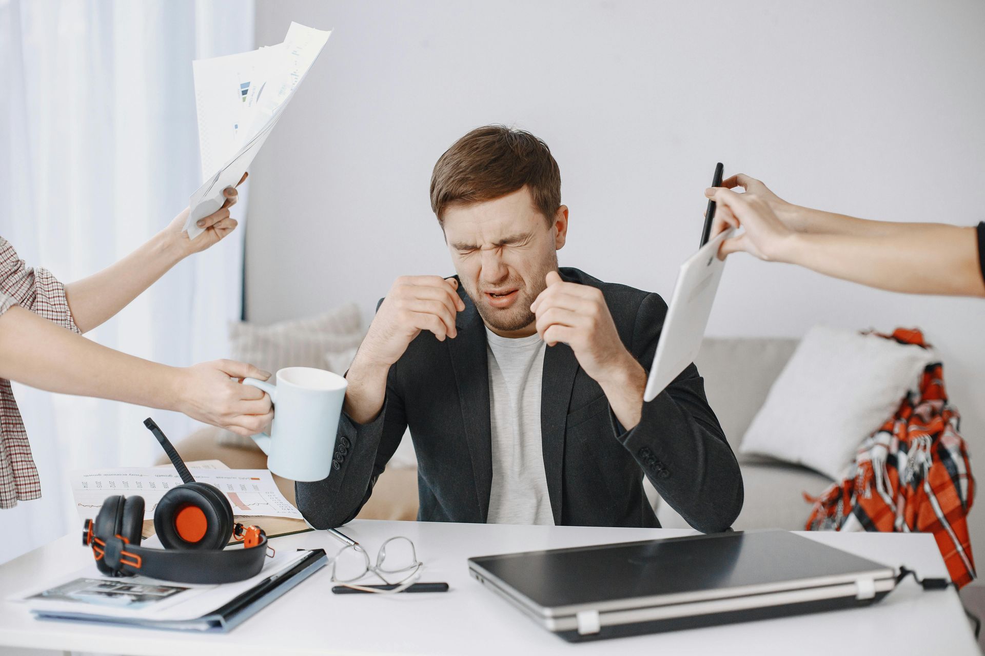 Man overwhelmed by demands, grabbing his face with papers, tablet, and coffee coming at him.