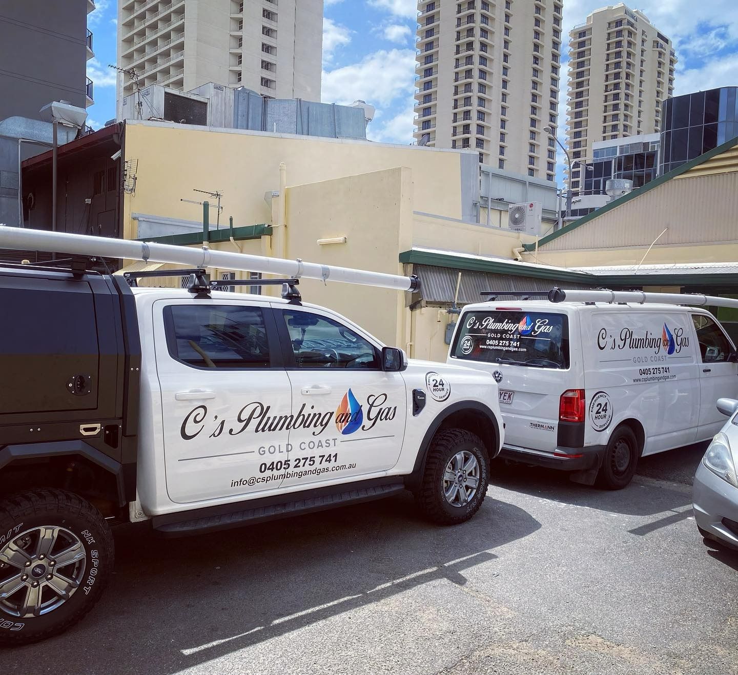Two branded plumbing vans with roof pipes parked outside in a city street with high-rise buildings.