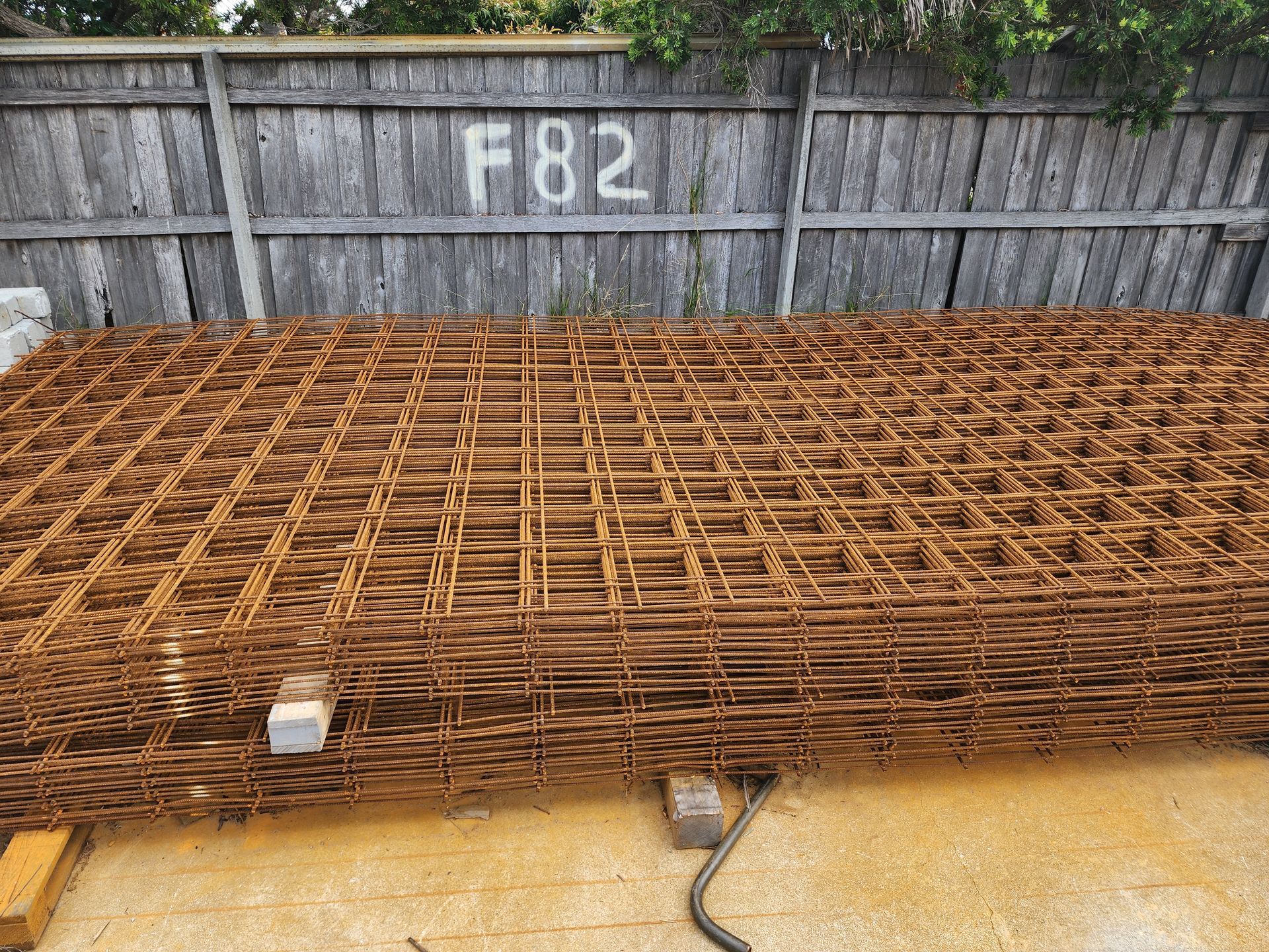 Rusty steel reinforcing mesh stacked on concrete blocks against a wooden fence. 