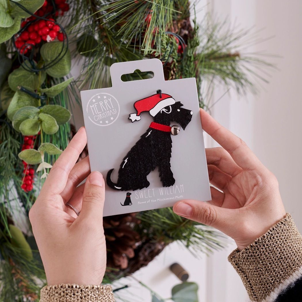 Kerstversiering voor een hond: een zwarte schnauzer met een kerstmuts en -halsband. Grijze kaart op hout met versieringen.