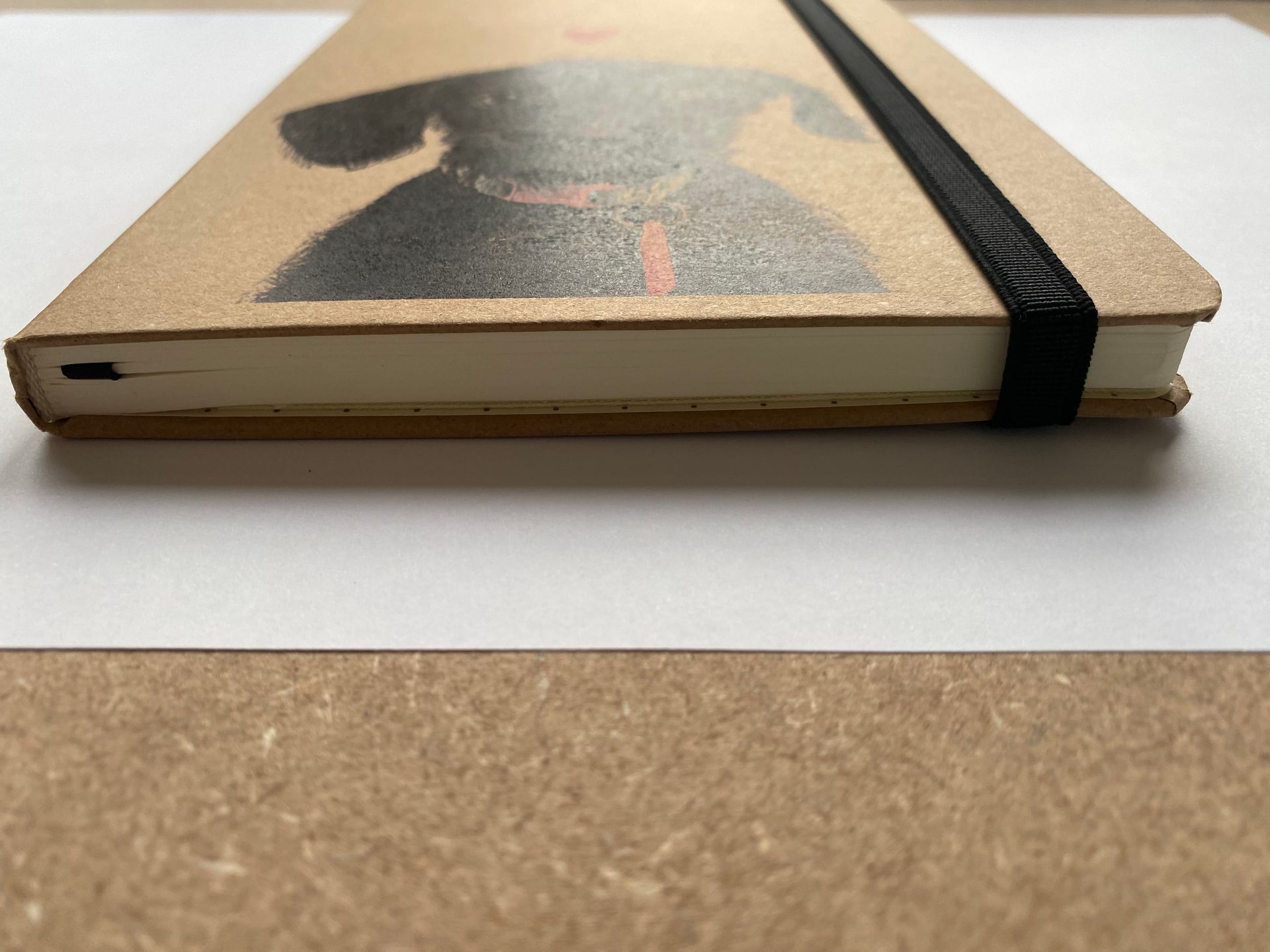 Notitieboekje met illustratie van een zwarte hond, roze hart en zwarte elastische band op een bruine kaft.