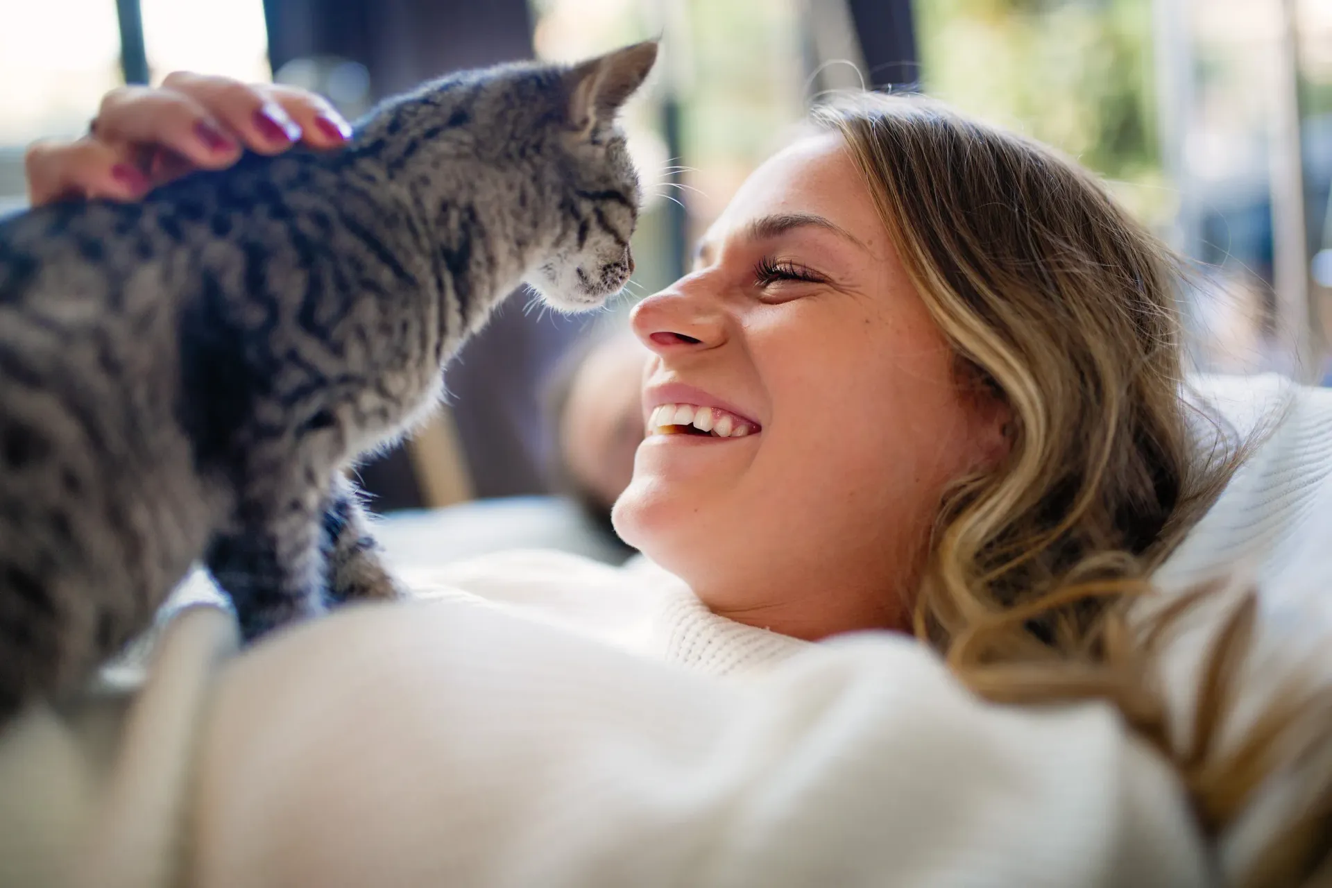 Woman smiling, petting a gray tabby cat lying on a white surface.