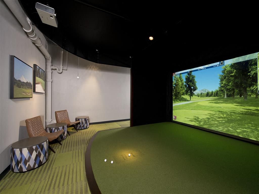 Indoor golf simulator room with a large screen, turf floor, and seating.
