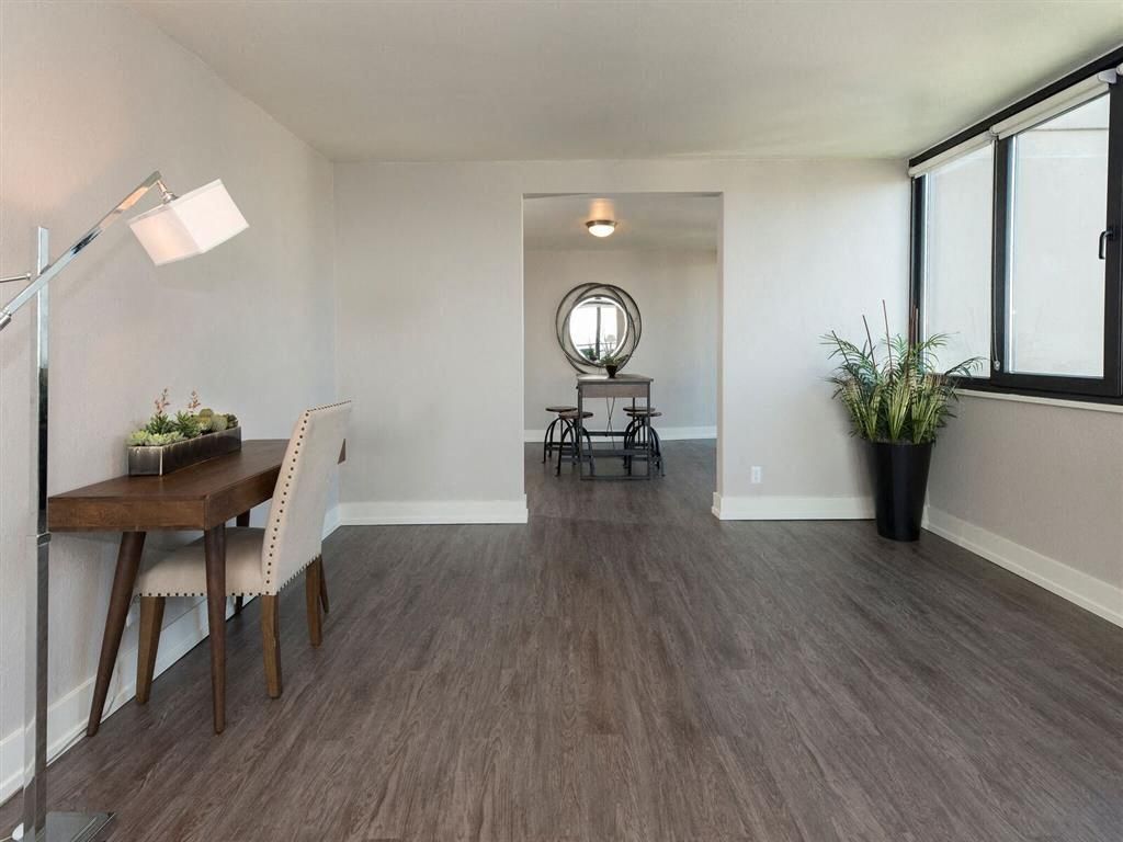 Interior living space with wood-like flooring, a desk and chair on the left, and large windows on the right.