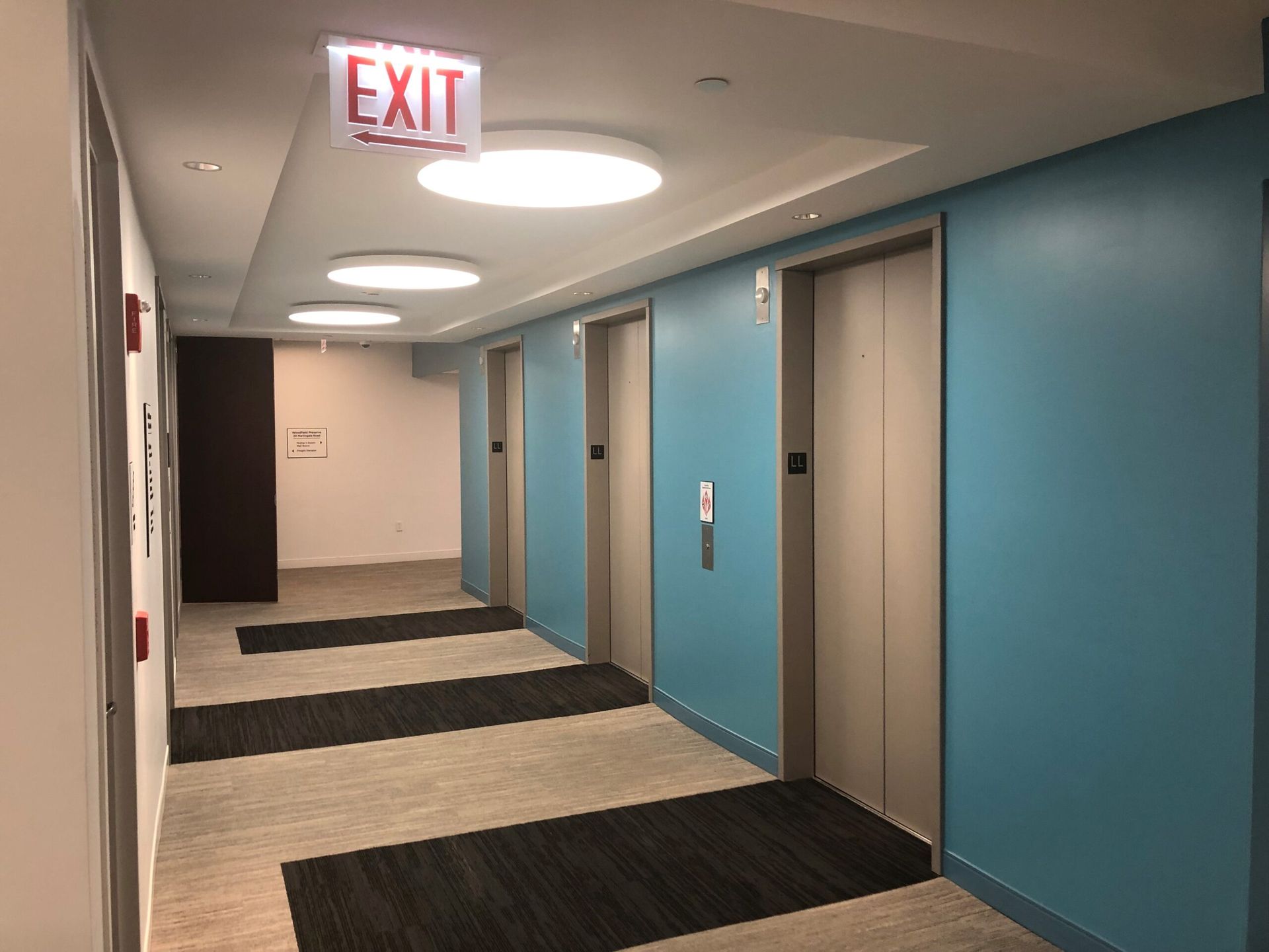 A hallway with elevators and an exit sign on the ceiling.