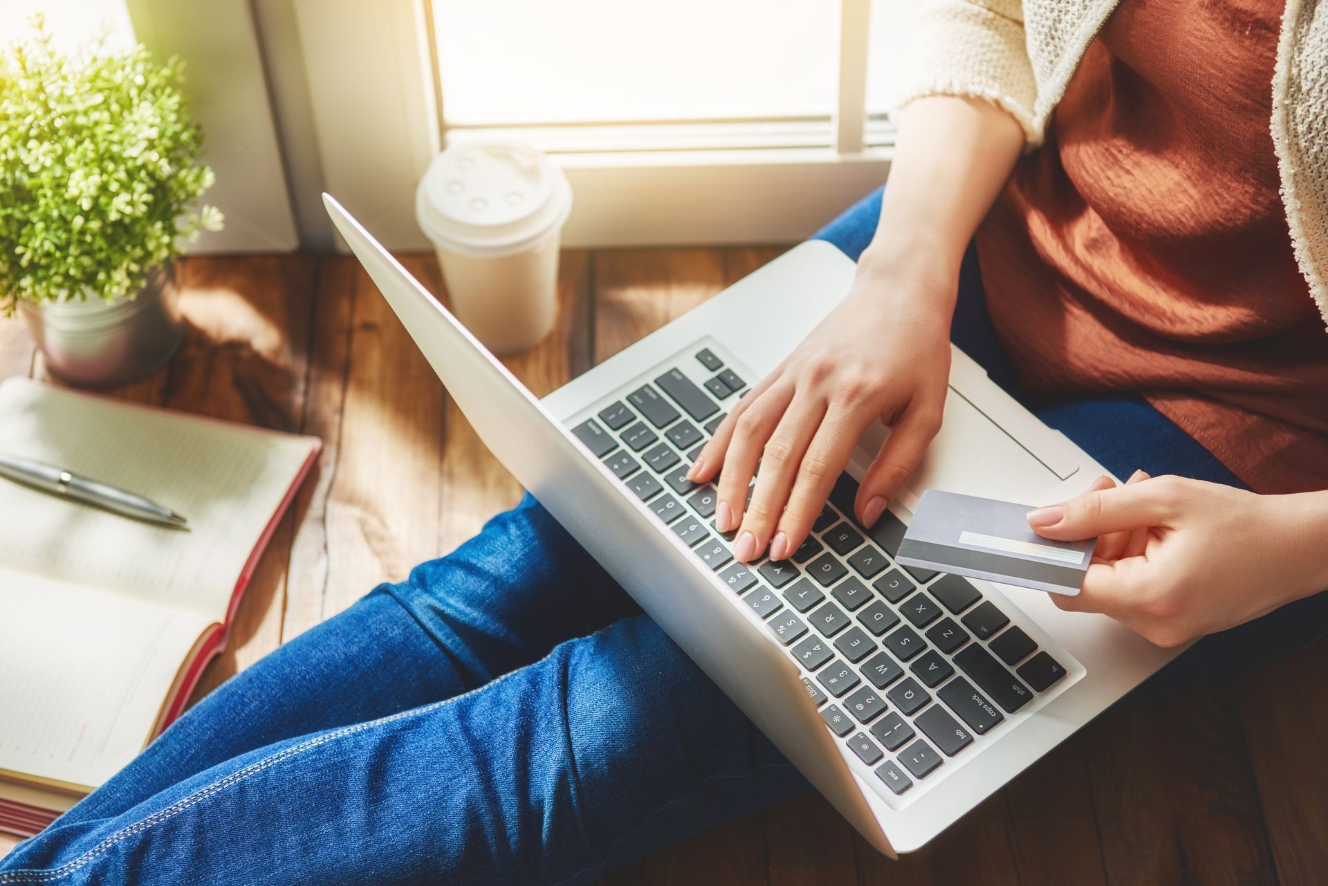 Person using a laptop on a couch beside a coffee cup and notebook in warm sunlight