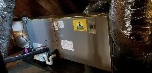 A large, rectangular HVAC unit sits in an attic. Ductwork and exposed wooden beams are visible.