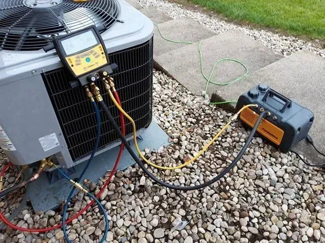 An HVAC technician working on an AC unit outdoors. Hoses are connected to gauges and a vacuum pump on a gravel surface.