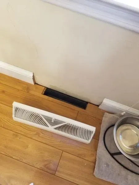 White and black floor air vents on a hardwood floor, one removed.