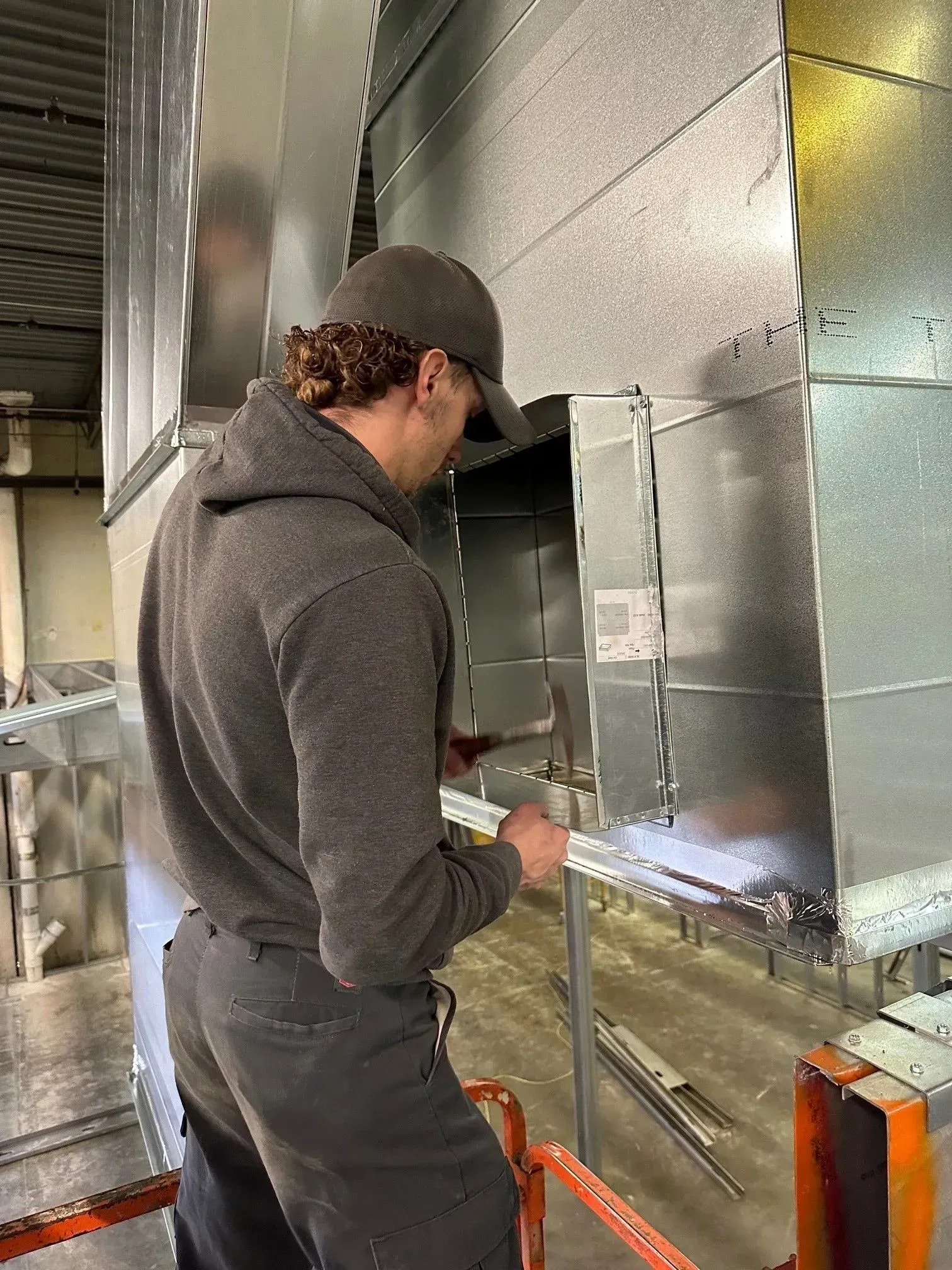 Man in gray hoodie working on metallic HVAC ductwork.