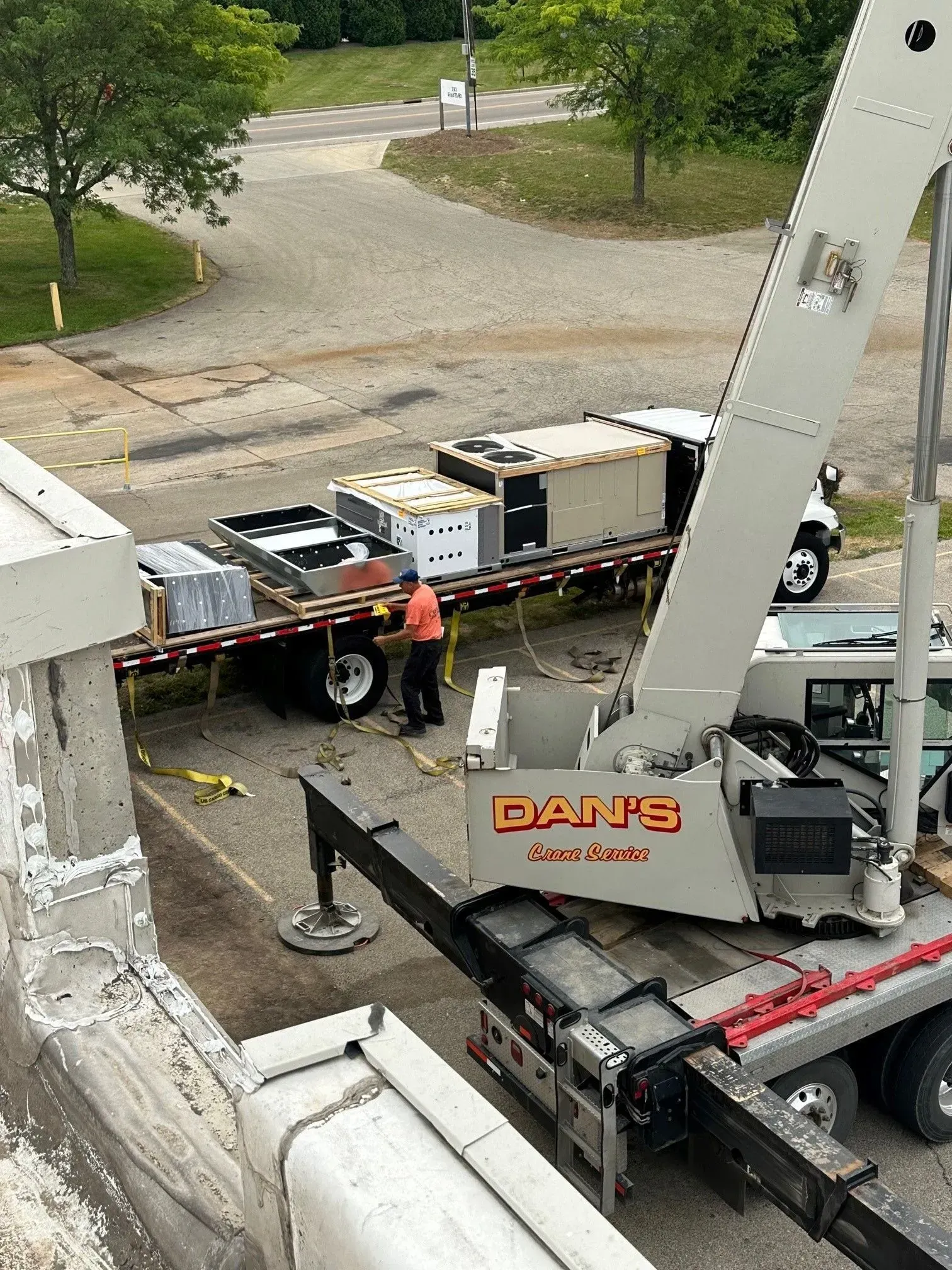 A crane lifts HVAC units off a flatbed truck. A worker in an orange shirt is present.