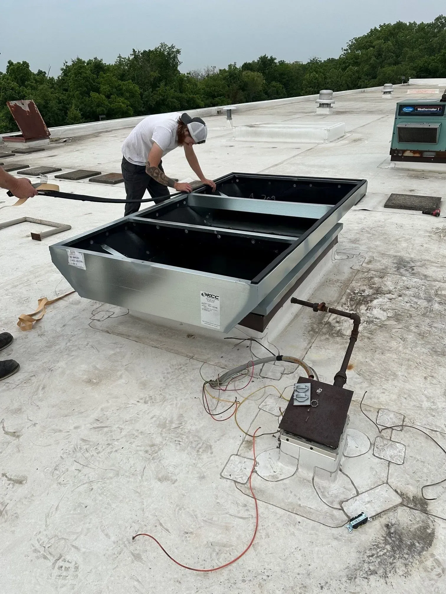 Person on rooftop installing HVAC equipment. A large, metal unit sits on a concrete surface. Trees are in the background.