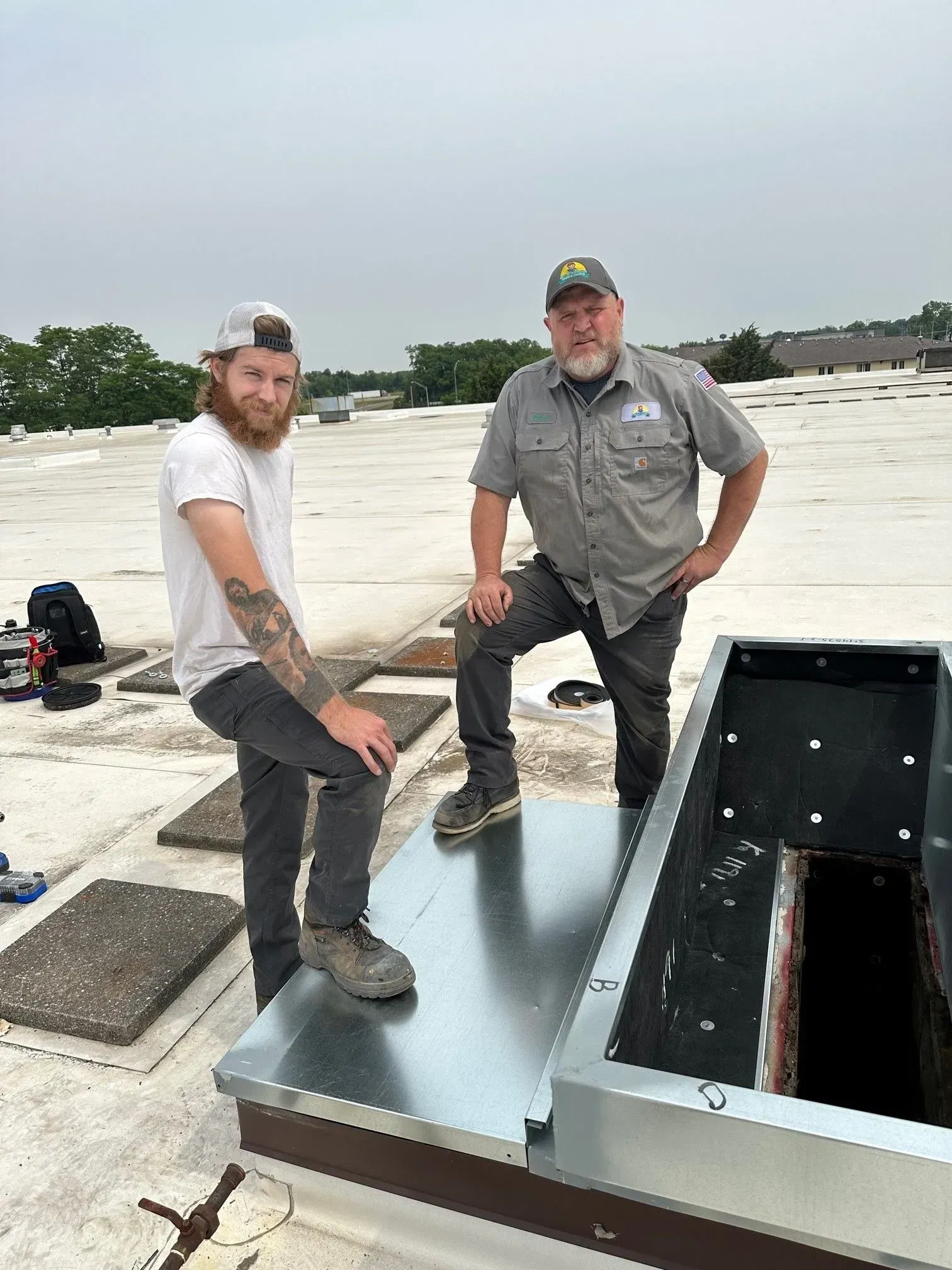 Two men on a roof near an open metal structure. One with beard, other has tattoos. Overcast day.
