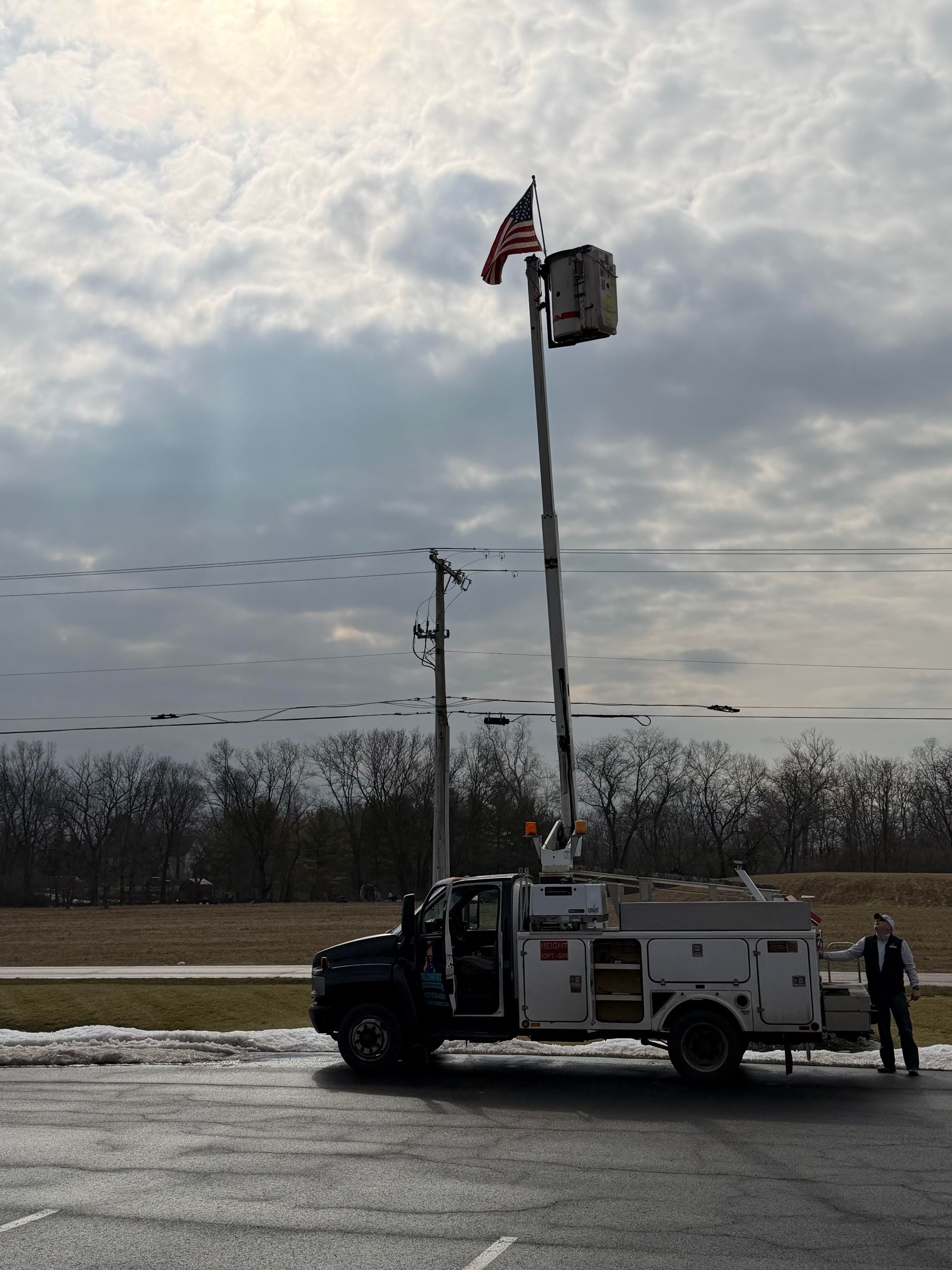 Utility truck with raised arm holding an American flag and a worker standing beside it. Cloudy sky in the background.