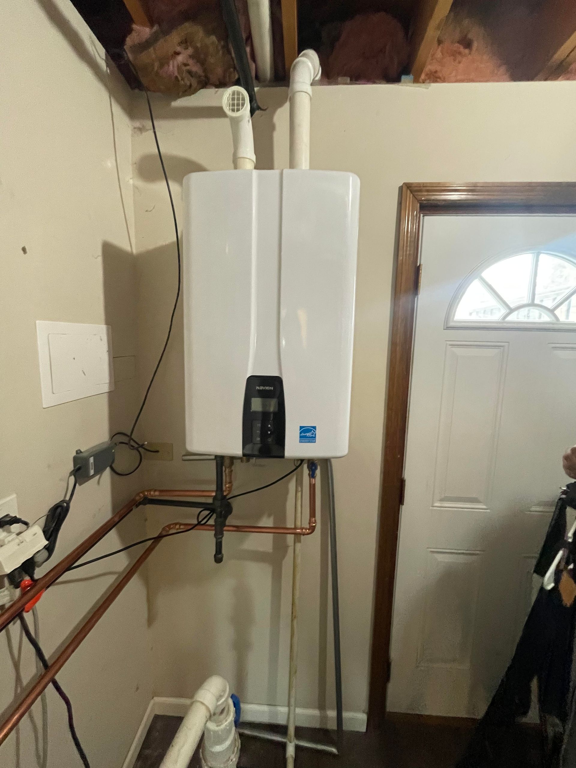White tankless water heater mounted on a stand. Copper pipes connect to the unit in a utility room. Door to the right.