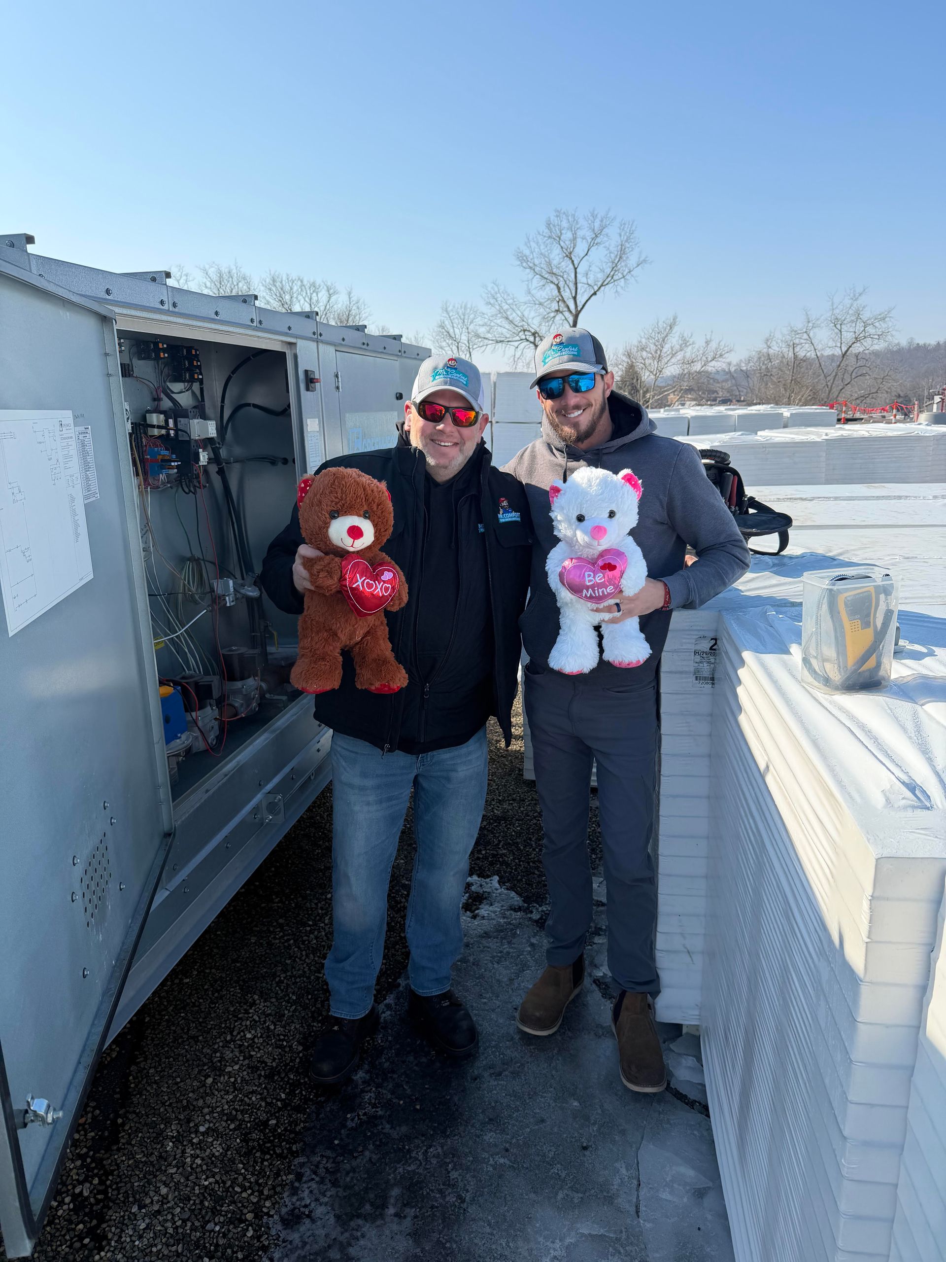Two men on a snowy rooftop holding Valentine's Day teddy bears near equipment; sunny day.