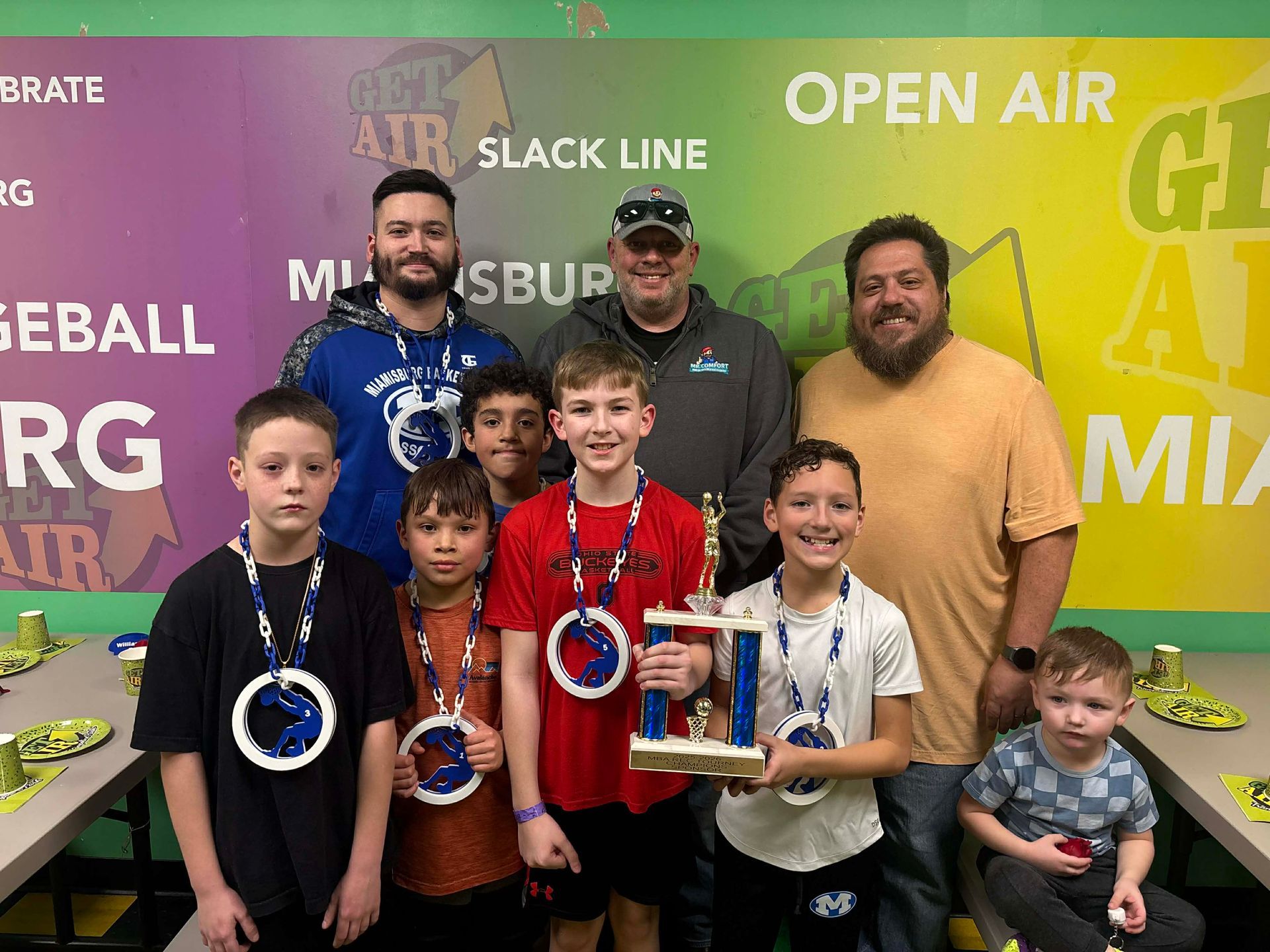 A group of kids with medals and adults holding a trophy, celebrating a win.