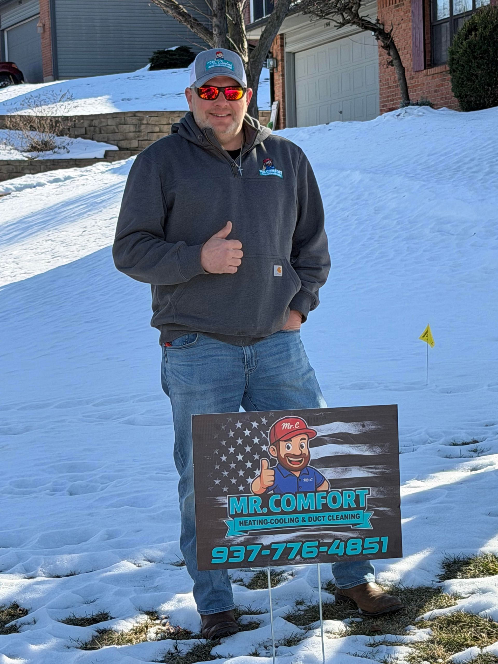 Man in a cap and hoodie gives a thumbs up next to a sign for Mr. Comfort HVAC in a snowy yard.