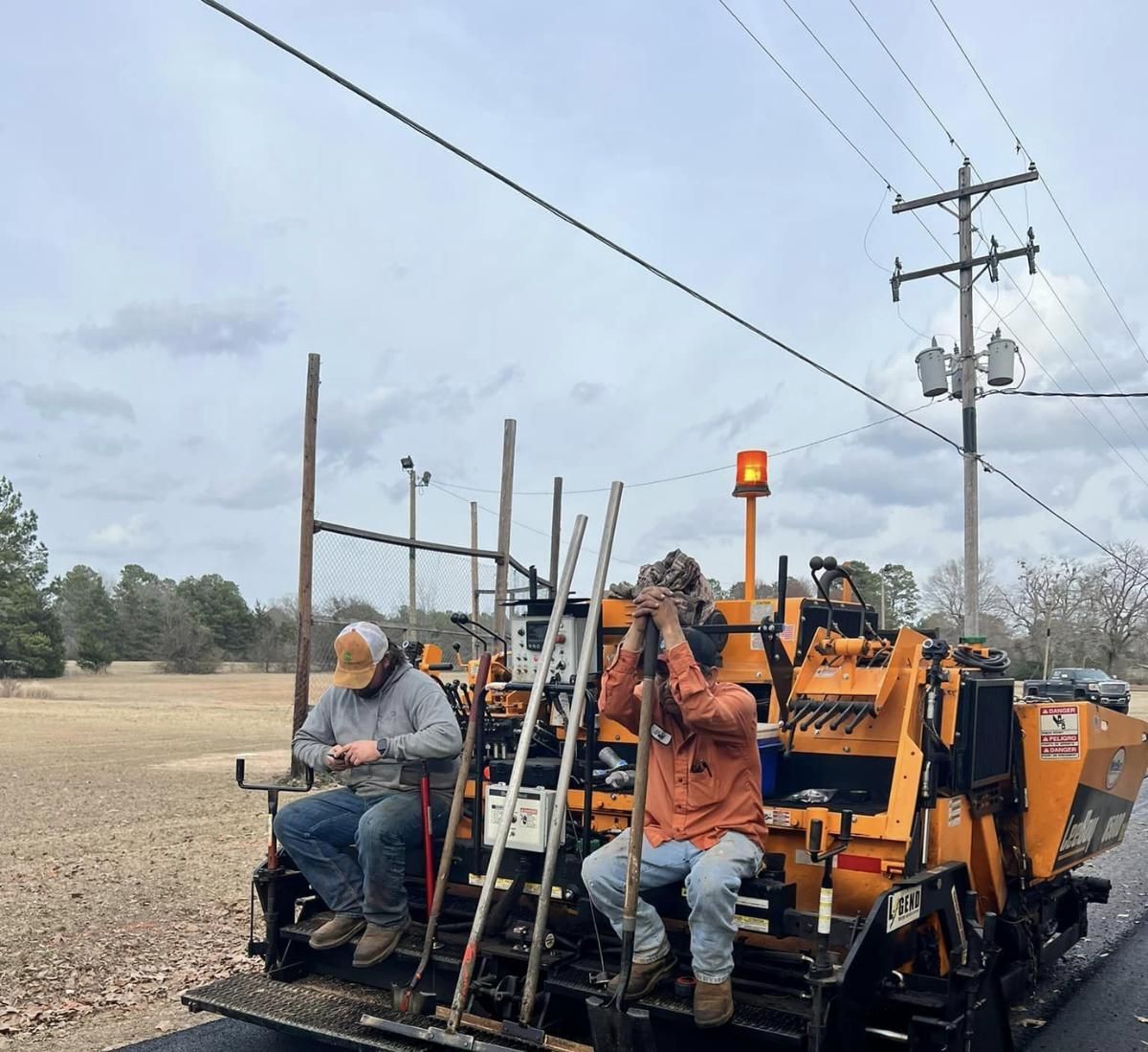 Two workers on an asphalt paving machine near power lines and a cloudy sky.