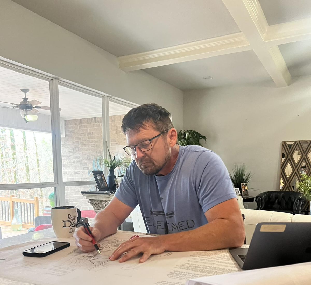 Man in glasses sketching on a blueprint at a table, with a mug, phone, and tablet nearby, next to a window.