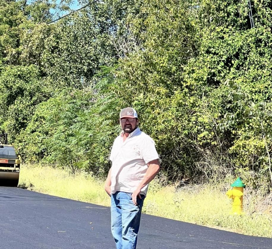 Man stands on new asphalt road next to a fire hydrant with a roller machine in the background and a lush green backdrop.