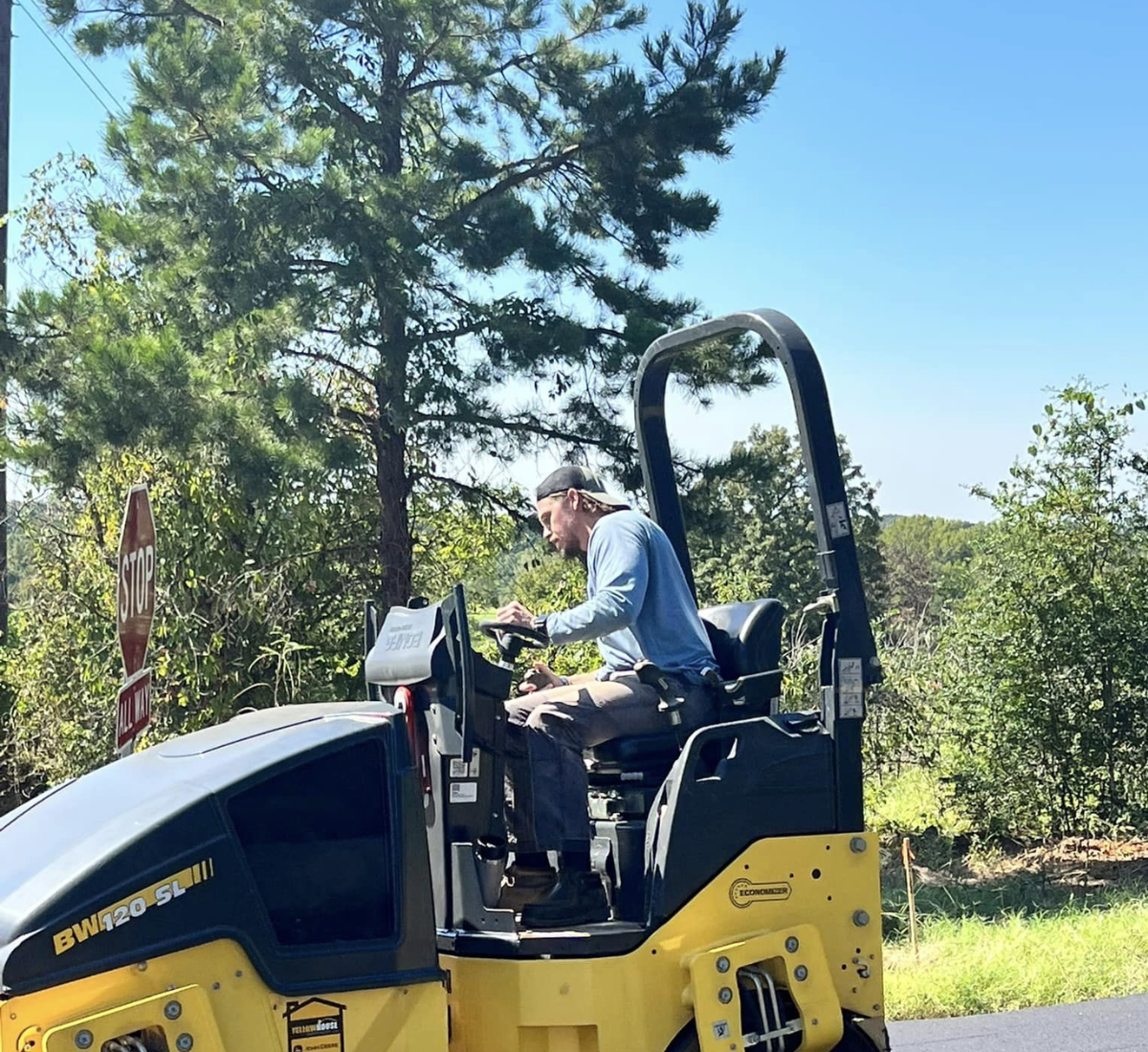 Man operating a yellow and black asphalt roller on a sunny day.