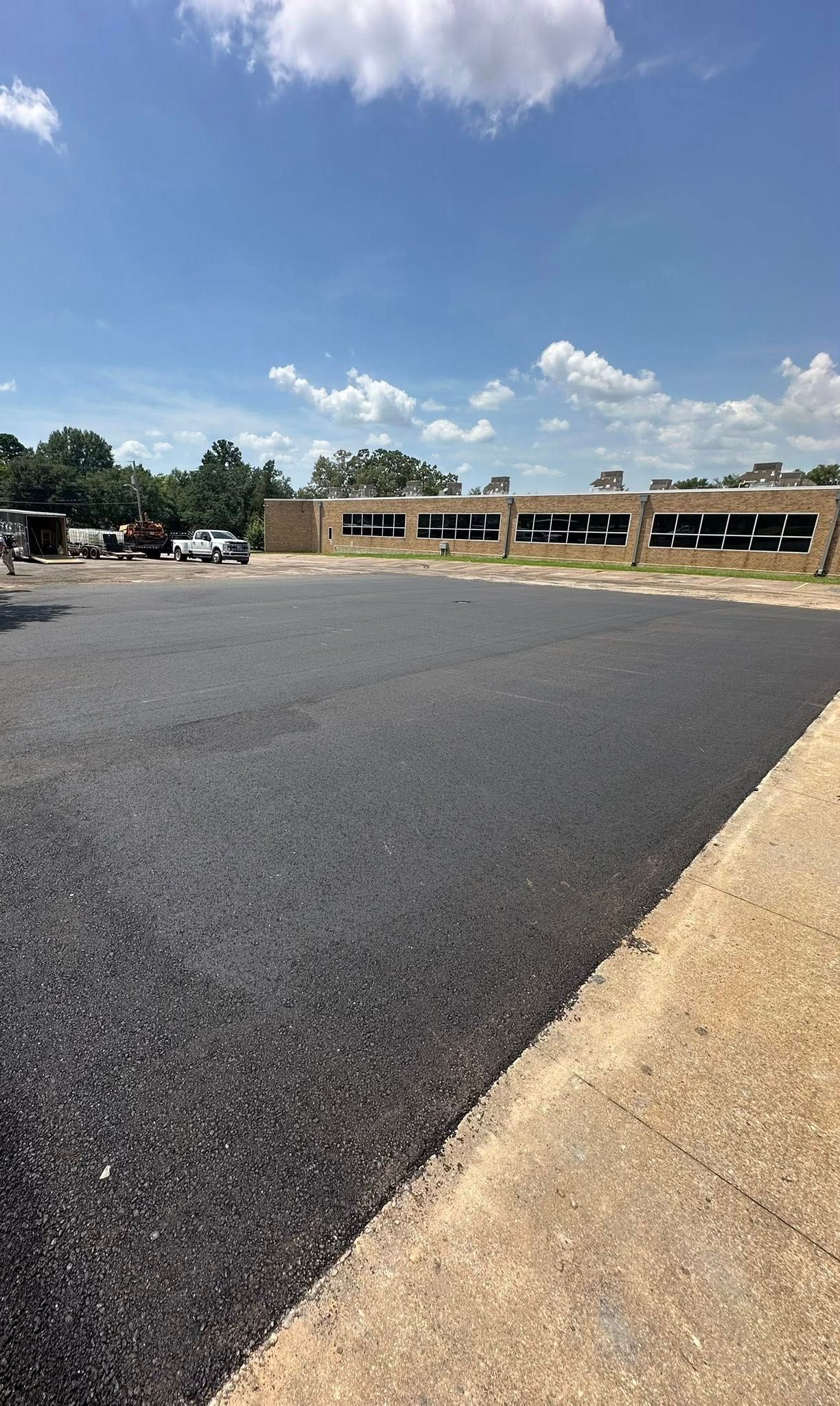Newly paved asphalt lot with a building and sky in the background, under a blue sky with some clouds.