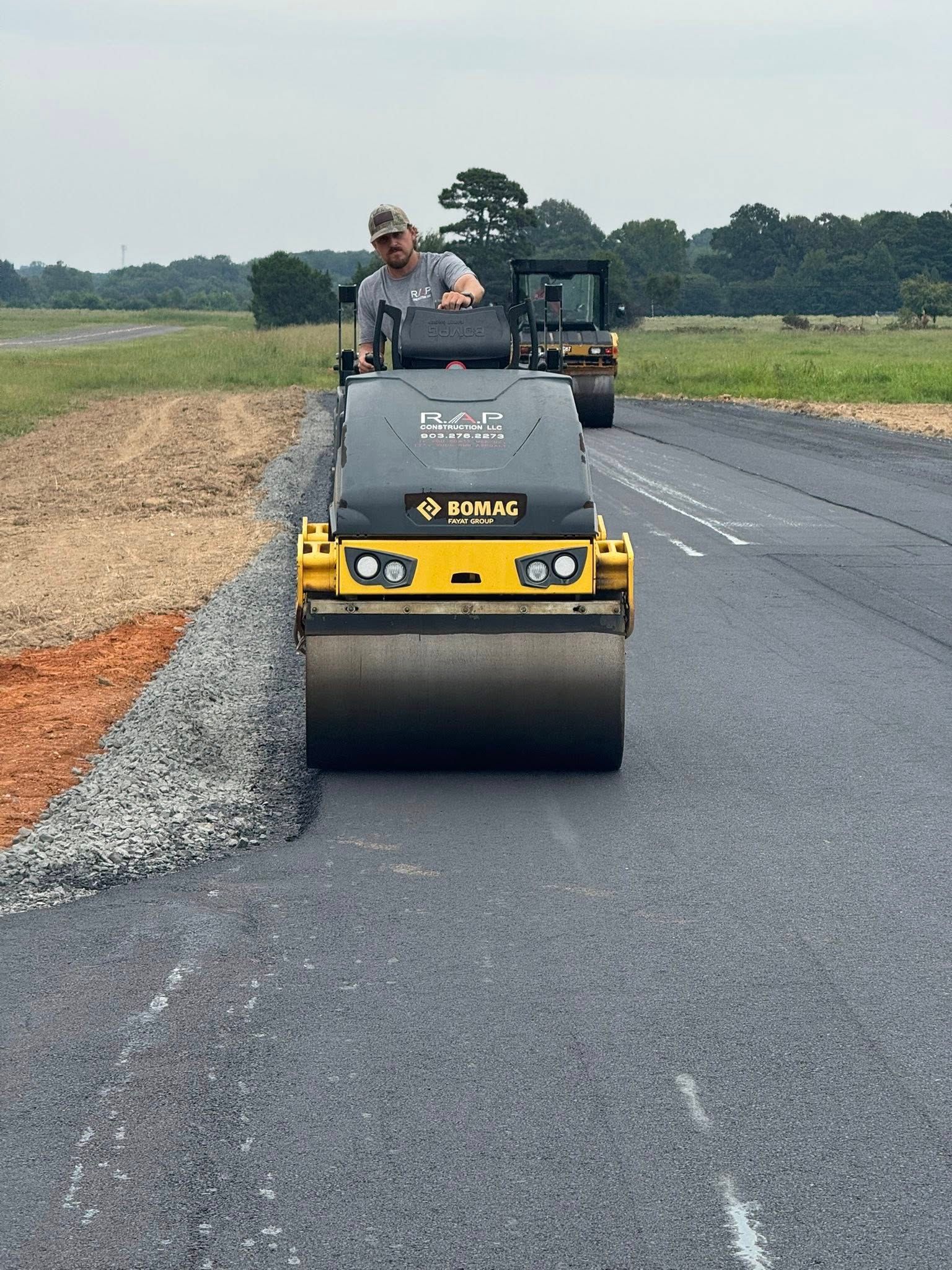 Man operating a yellow road roller on a newly paved road; another machine in background.