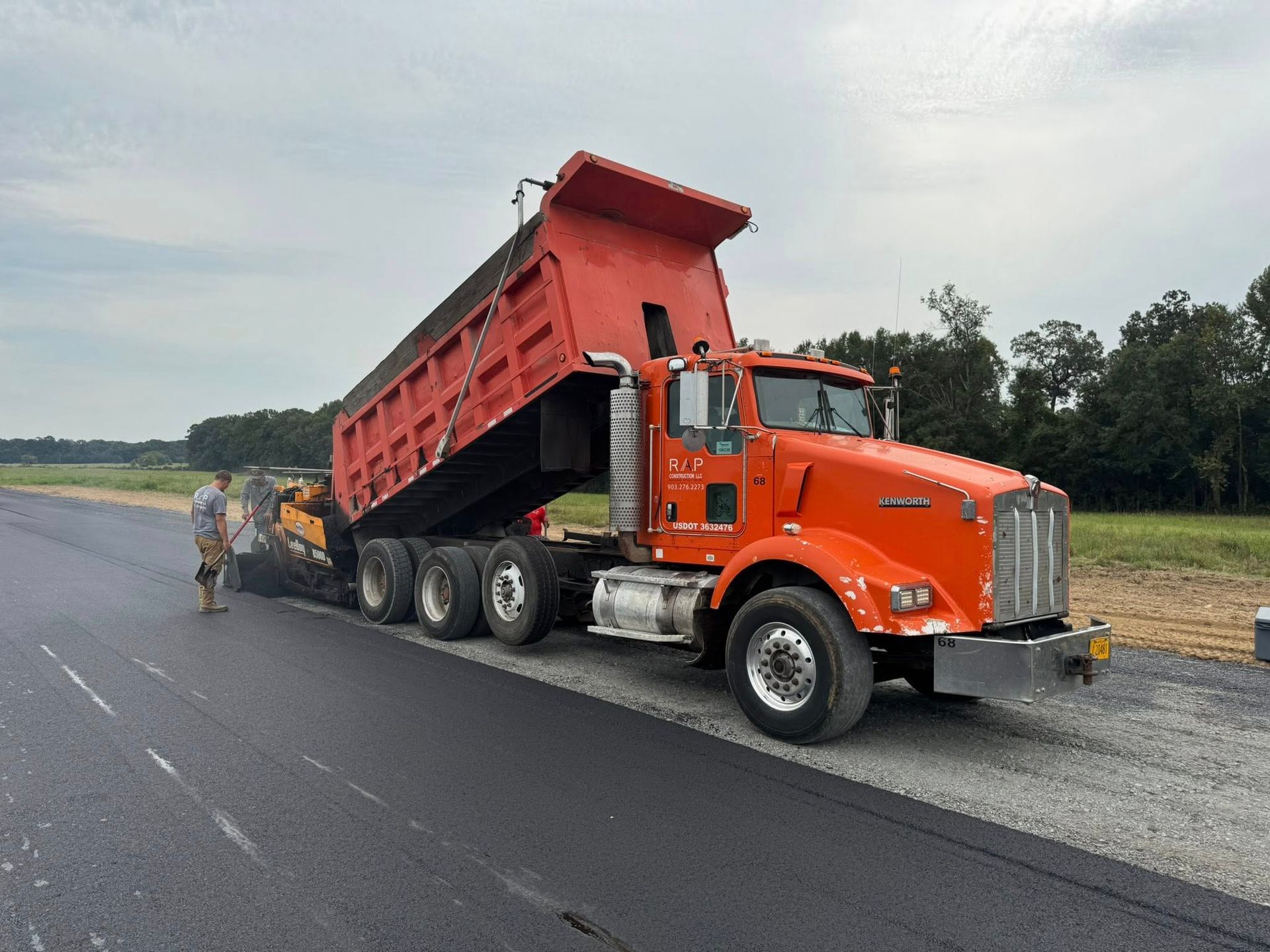 Orange dump truck unloading asphalt into a paver on a road construction site.