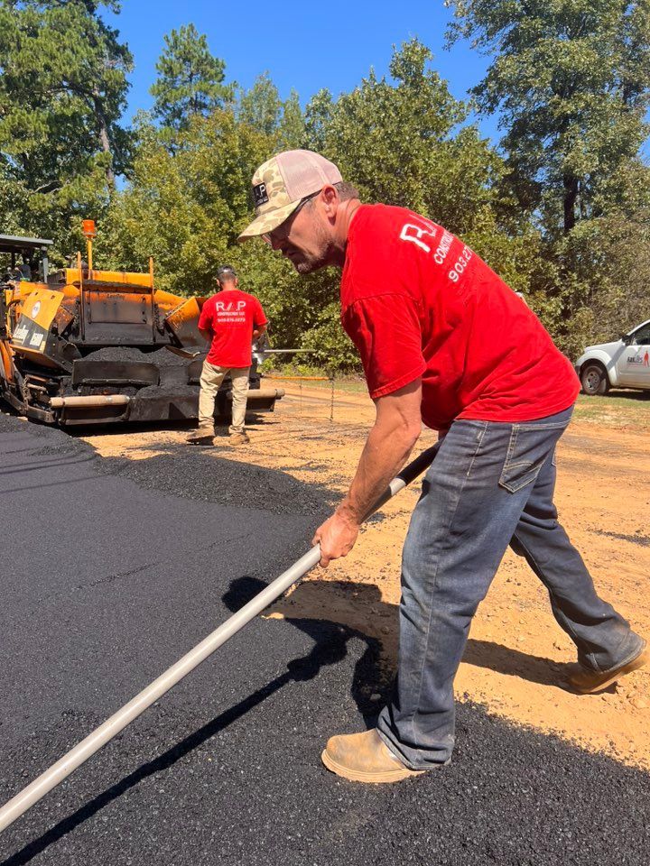 Man using a rake to smooth fresh asphalt on a road. Another worker and paving equipment are in the background.