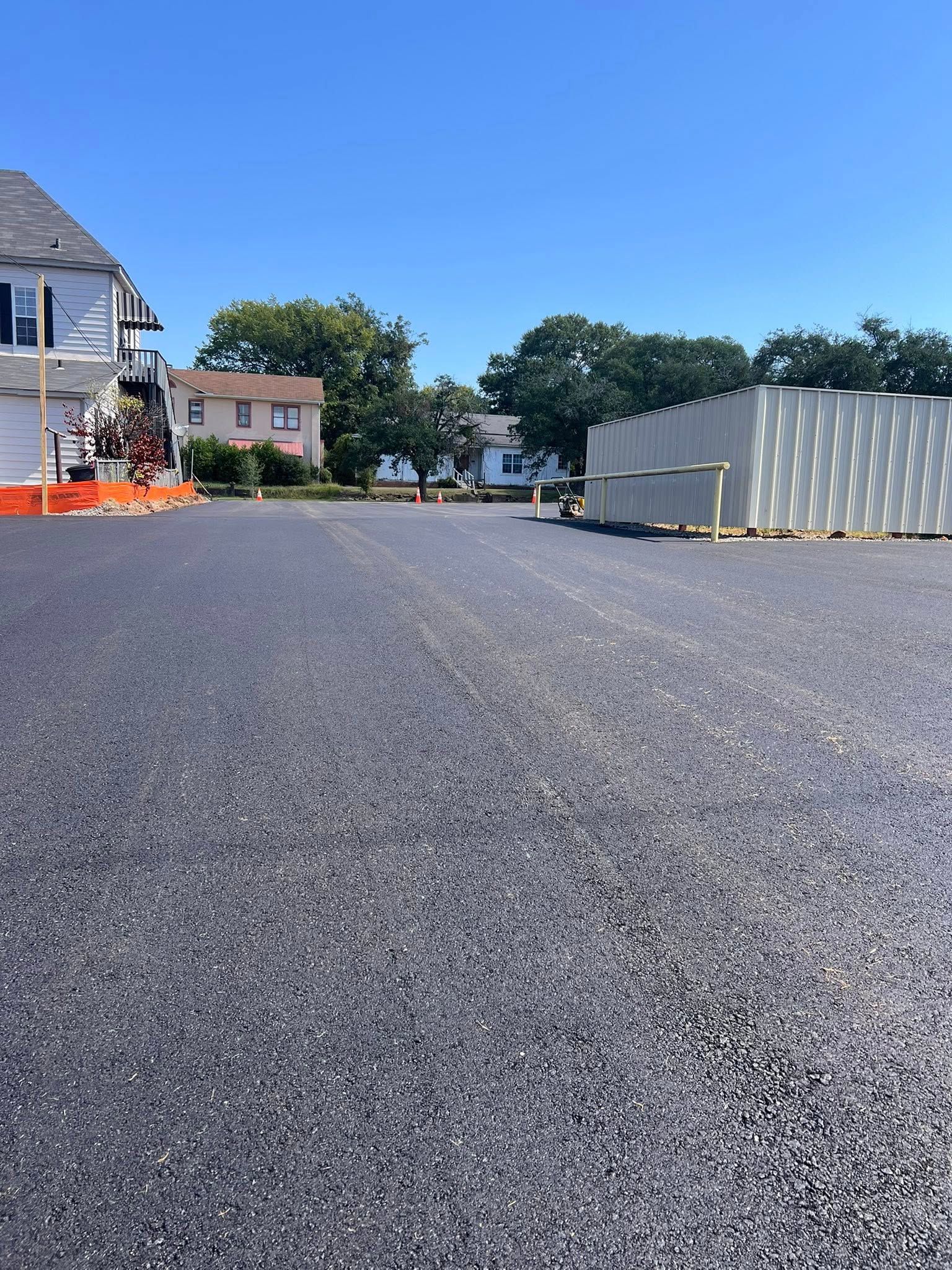 Newly paved asphalt parking lot on a sunny day. Buildings and trees in the background under a blue sky.