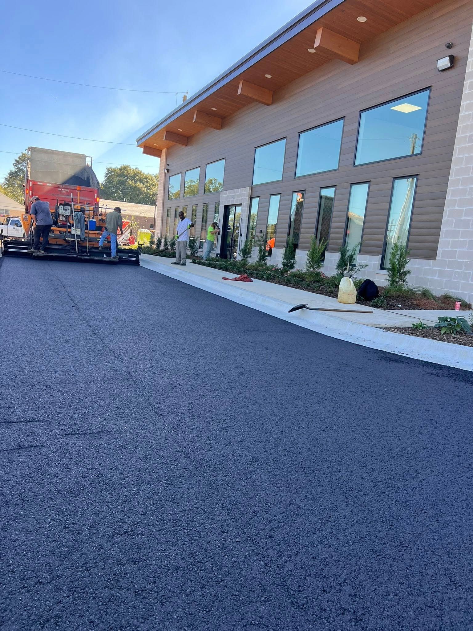 Paving crew laying asphalt in front of a modern building with large windows.