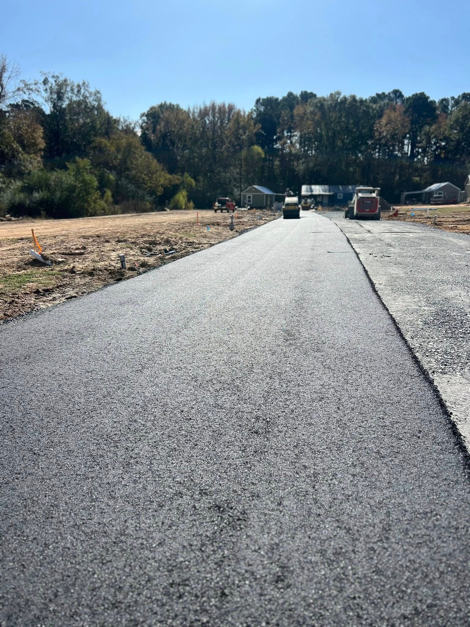 Newly paved asphalt road on construction site. Gray, gravelly surface extends toward horizon; trees in background.