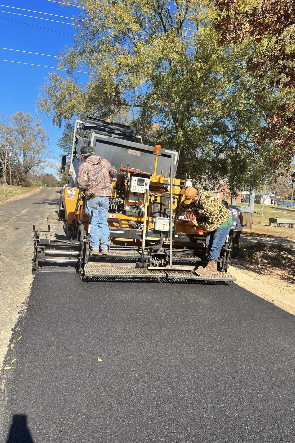 Road paving machine laying asphalt on a street. Two workers in camo adjust the machine under a sunny sky.