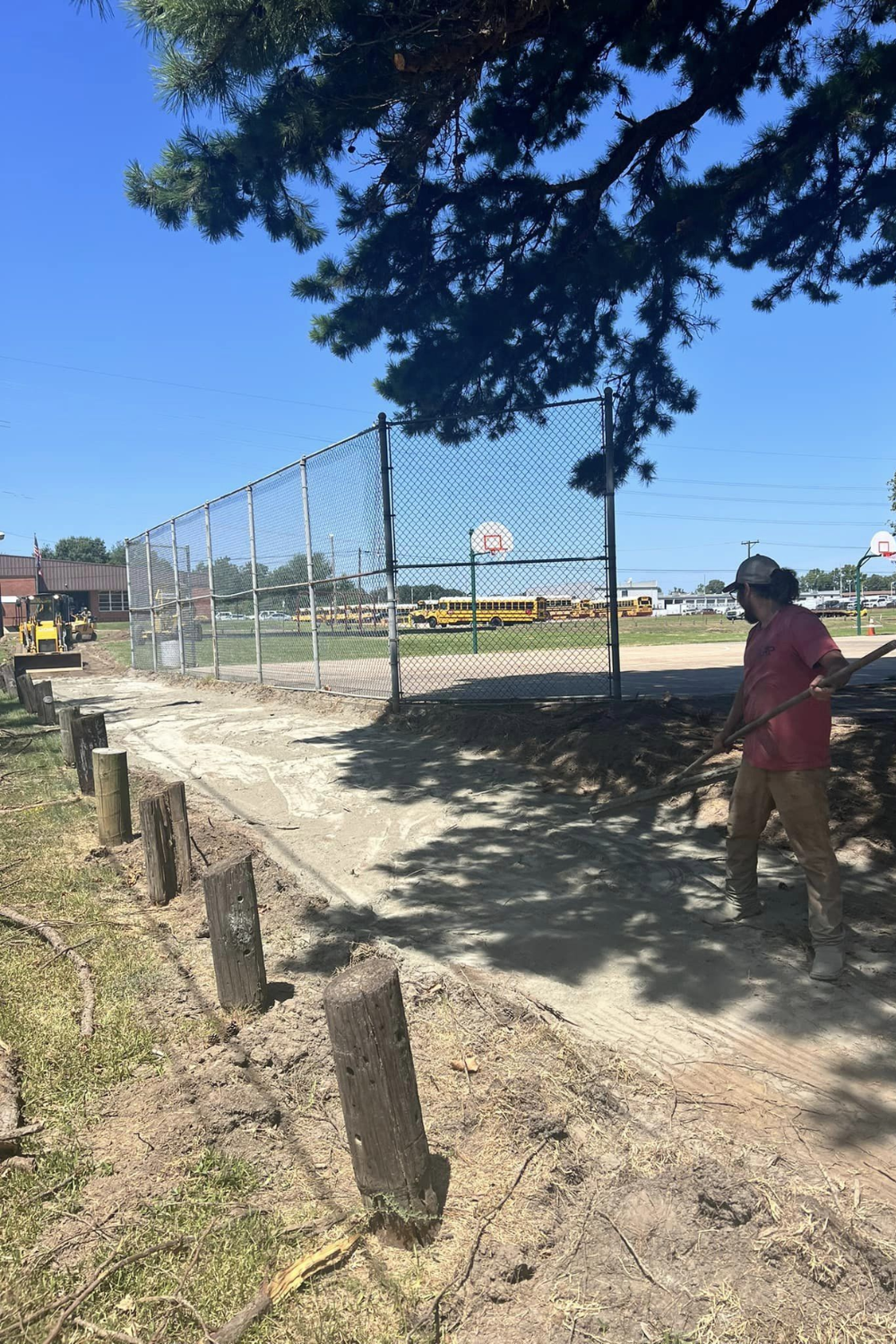A person sweeps a dirt path next to a fence and a basketball hoop on a sunny day.