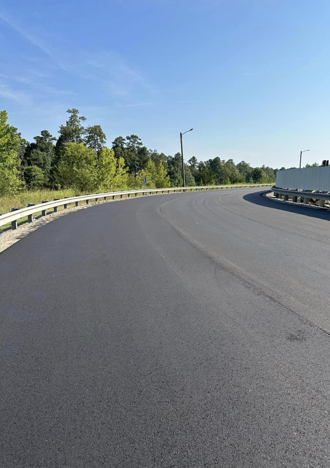 Newly paved road curves along a tree line, with a blue sky overhead.