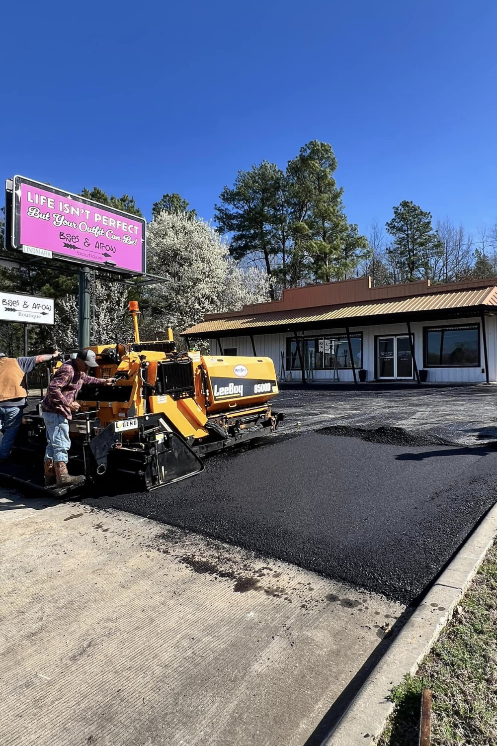 Asphalt paving a parking lot; worker operates machine. Building in background, blue sky.