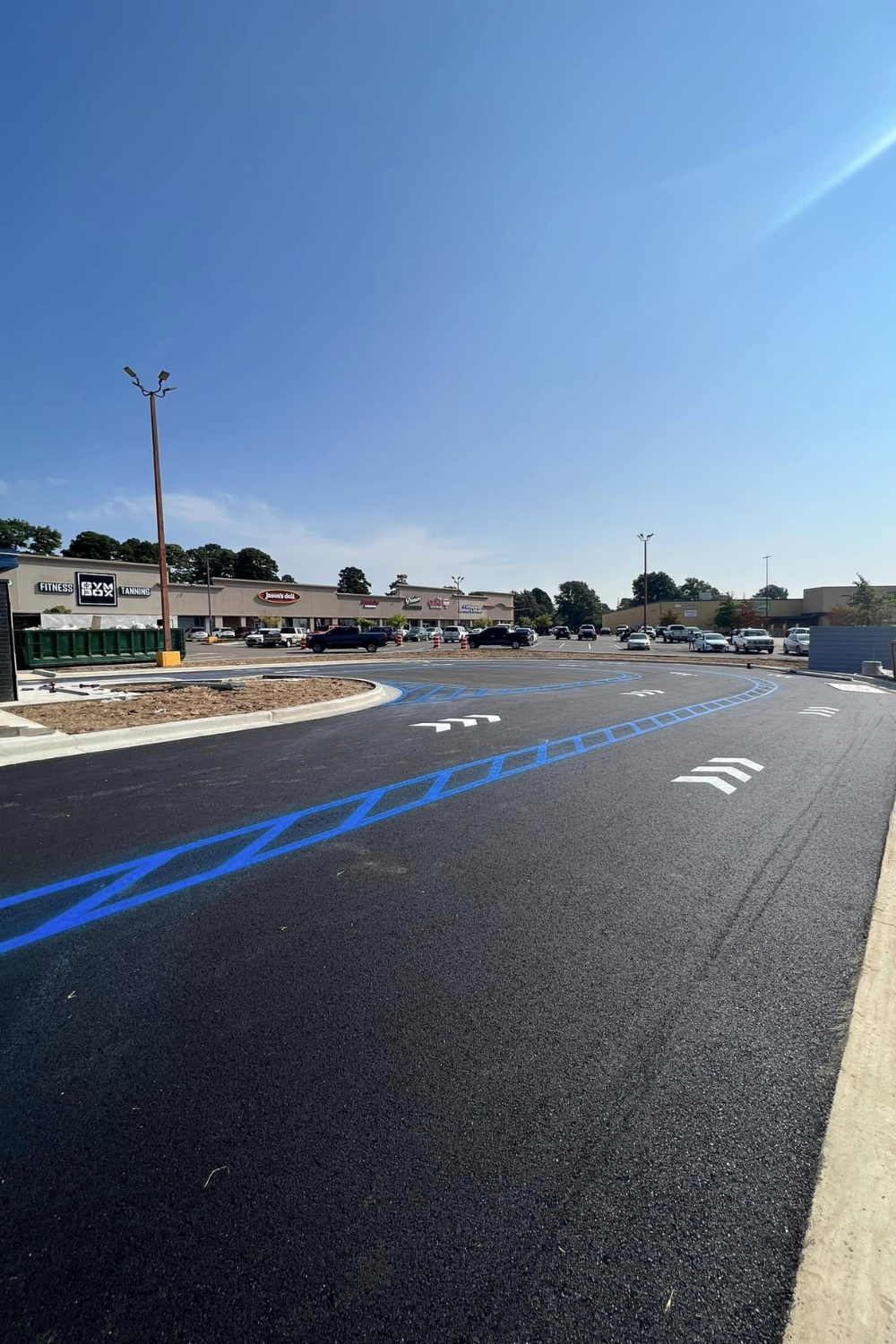 Newly paved asphalt lot with blue line markings and white directional arrows. Shopping center in the background.